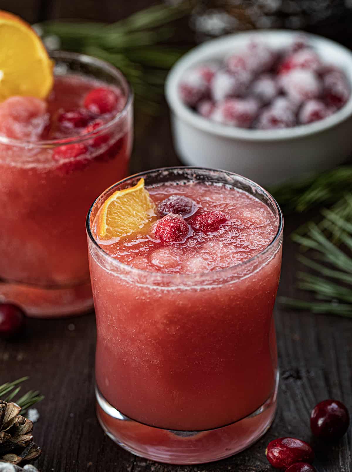 Christmas Slush Mocktail is a refreshing and festive beverage that can be enjoyed by all ages. It's made with a mixture of cranberry juice, orange juice, and lemonade concentrate, which is then frozen and served topped with cherry lemon-lime soda. Glass of Christmas Slush with an Orange Garnish and Sugared Cranberries.