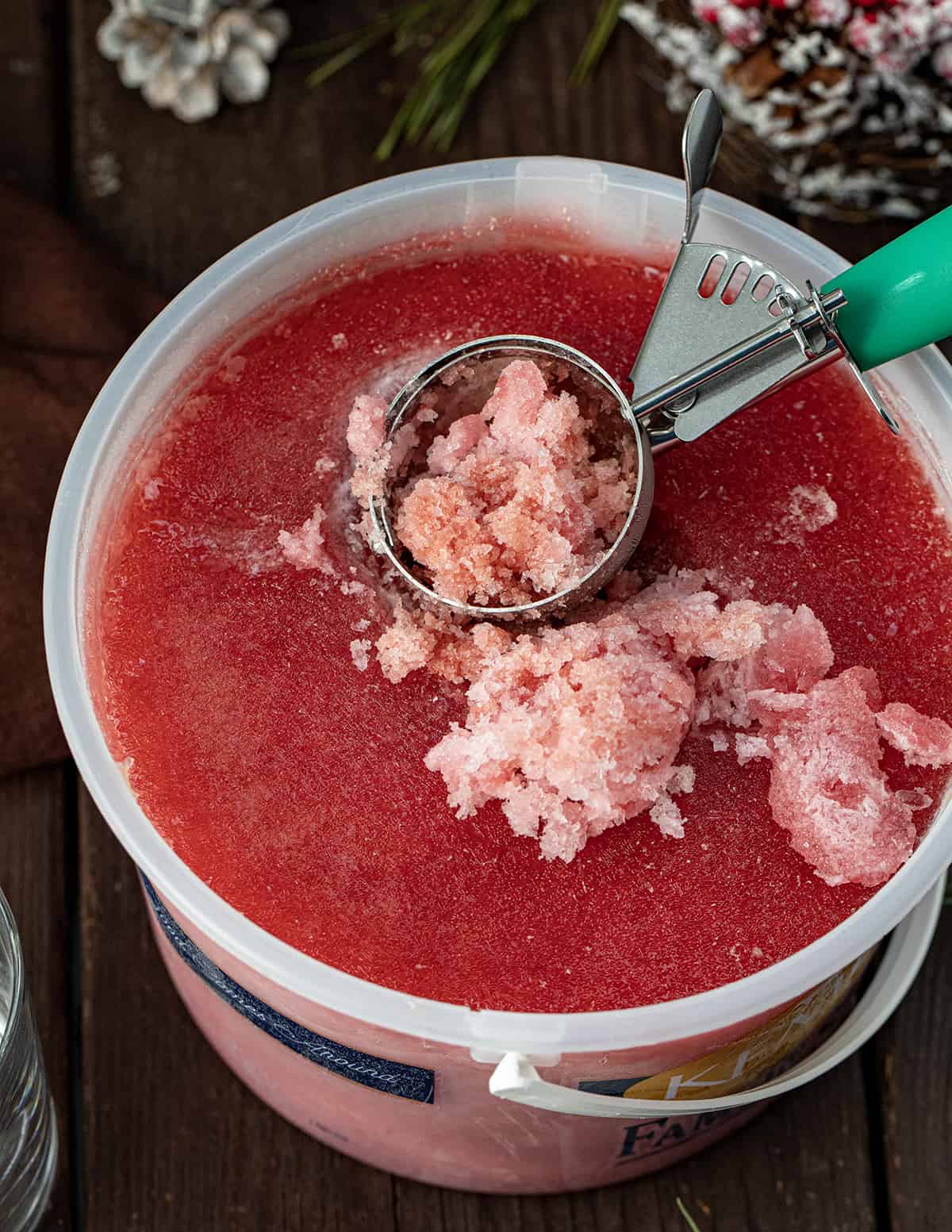 Christmas Slush Mocktail is a refreshing and festive beverage that can be enjoyed by all ages. It's made with a mixture of cranberry juice, orange juice, and lemonade concentrate, which is then frozen and served topped with cherry lemon-lime soda. Bucket of Christmas Slush with Some Scooped Out.