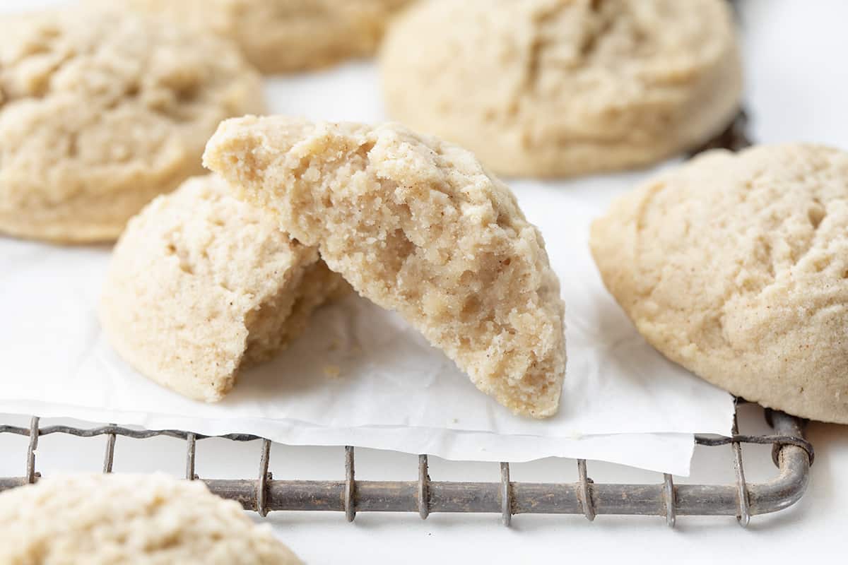 Cinnamon Butter Cookies are tender and crumbly cookies that combine the warm flavor of cinnamon and the richness of butter. The sweet buttery flakiness paired with a subtle cinnamon kick is a delectable combination. It's the perfect addition to any cookie exchange, holiday gathering, or Tuesday! Cinnamon Butter Cookies on a Cooling Rack with Parchment and a Cookie Halved Showing Inside Texture.