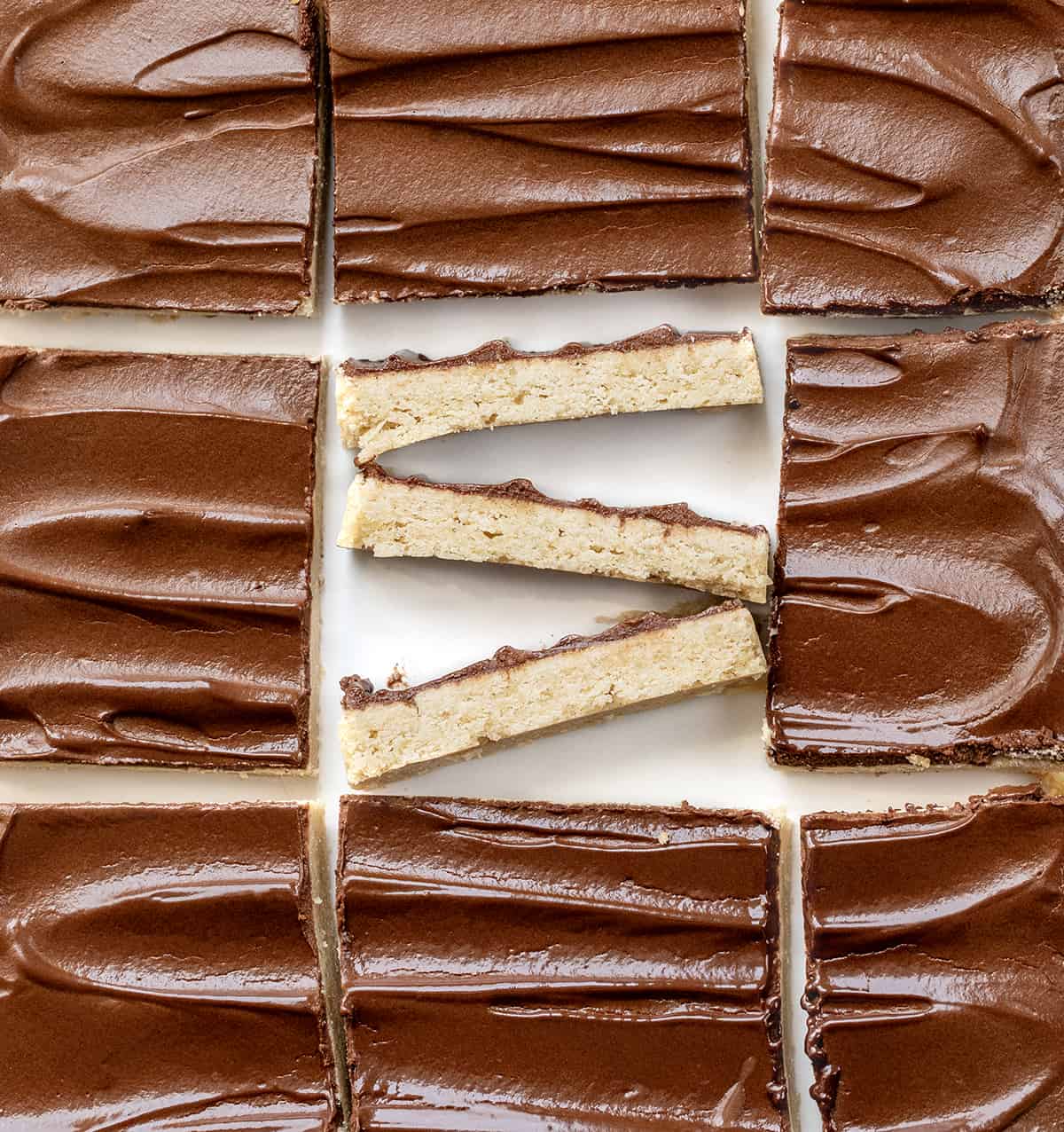 Cinnamon Shortbread Bars start with a buttery and crumbly shortbread cookie base flavored with cinnamon, topped with a whipped chocolate ganache. The ganache adds a rich and decadent layer that complements the cinnamon in the shortbread. Cinnamon Shortbread Bars on a White Counter with the Center Bar Cut up and Put on its Side.