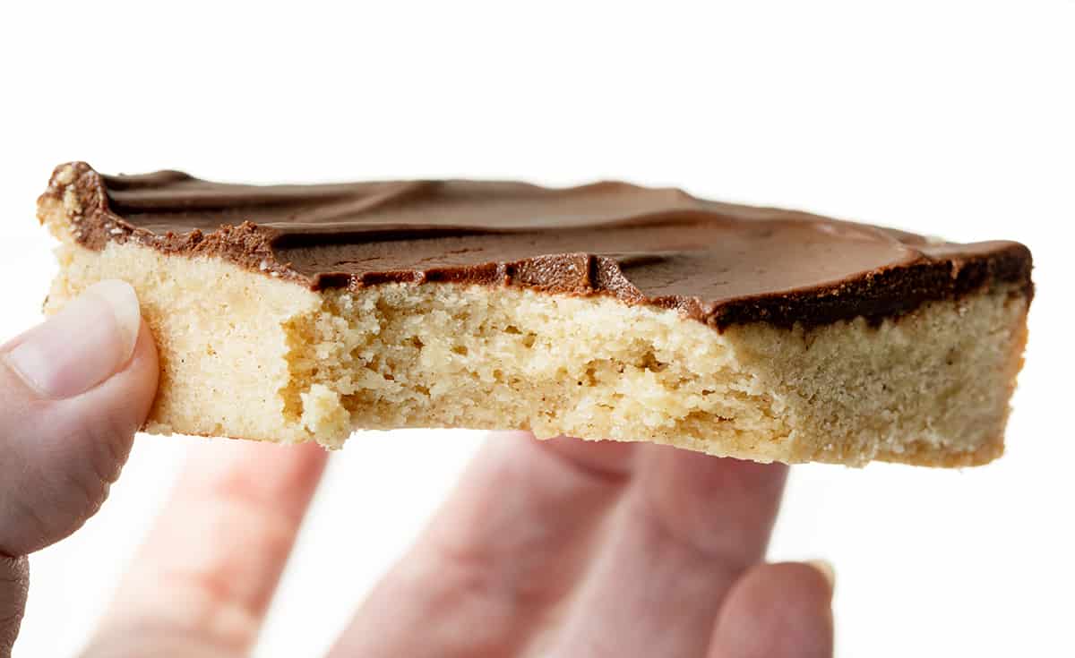Cinnamon Shortbread Bars start with a buttery and crumbly shortbread cookie base flavored with cinnamon, topped with a whipped chocolate ganache. The ganache adds a rich and decadent layer that complements the cinnamon in the shortbread. Hold holding a Bit Into Cinnamon Shortbread Bar.