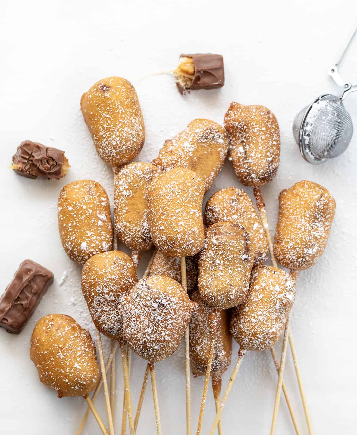Deep Fried Snickers are fun and indulgent treats that combine the rich and gooey texture of a Snickers bar with a deep-fried golden exterior. Then, they are dusted with confectioners' sugar for beauty, but that is definitely an optional step! Serving them on a stick is a playful twist that adds to the overall experience, bringing a touch of that State Fair-like charm! Several Deep Fried Snickers Still on the Skewer on a White Counter.