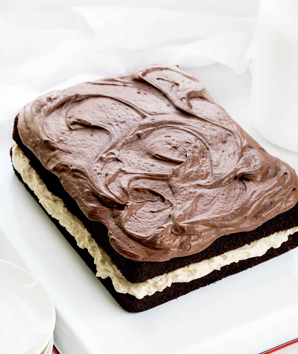 Ding Dong Sheet Cake is a decadent dessert with two layers of chocolate cake sandwiched together with a creamy filling and topped with chocolate frosting. The finished cake resembles a giant, elevated version of the popular chocolate-coated snack cake known as a "Ding Dong." It makes for a stunning and indulgent treat that is sure to delight chocolate lovers! Ding Dong Sheet Cake from A bit Higher Up Showing it Topped with Chocolate Ermine on a White Counter.