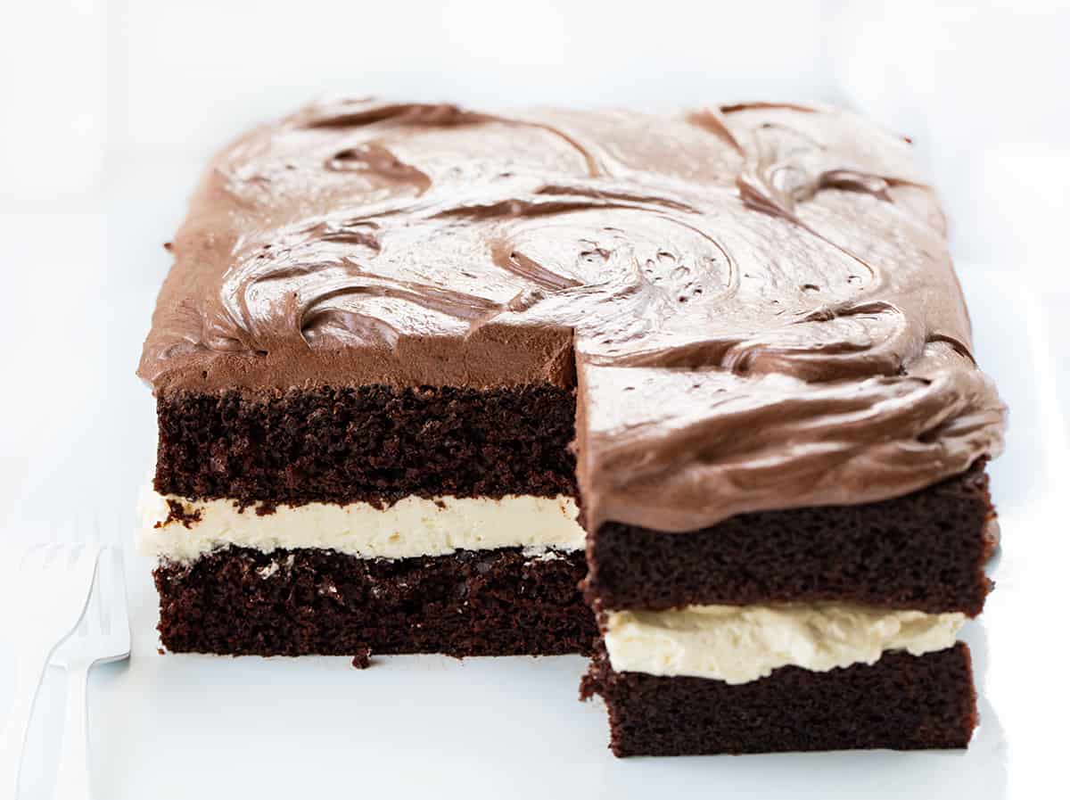 Ding Dong Sheet Cake is a decadent dessert with two layers of chocolate cake sandwiched together with a creamy filling and topped with chocolate frosting. The finished cake resembles a giant, elevated version of the popular chocolate-coated snack cake known as a "Ding Dong." It makes for a stunning and indulgent treat that is sure to delight chocolate lovers! Ding Dong Sheet Cake with Pieces Cut Out Showing Inside on a White Counter.