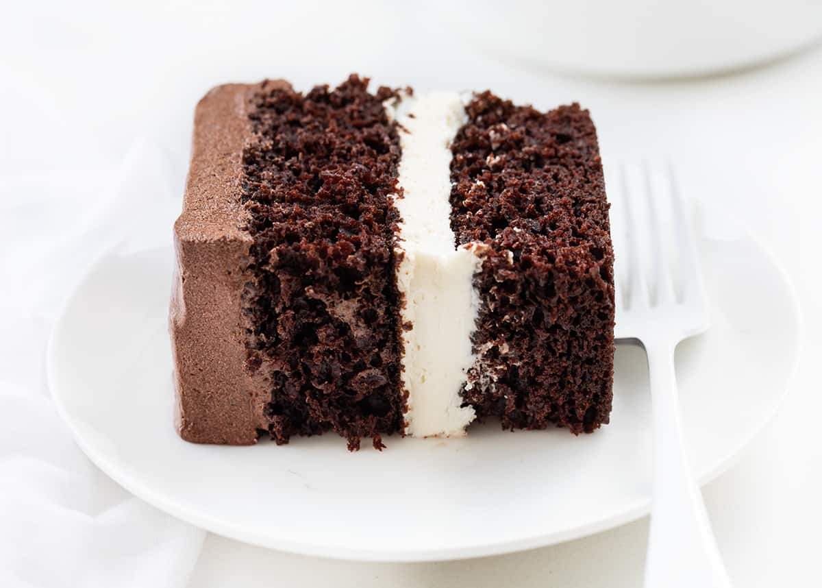Ding Dong Sheet Cake is a decadent dessert with two layers of chocolate cake sandwiched together with a creamy filling and topped with chocolate frosting. The finished cake resembles a giant, elevated version of the popular chocolate-coated snack cake known as a "Ding Dong." It makes for a stunning and indulgent treat that is sure to delight chocolate lovers! Piece of Ding Dong Sheet Cake on a White Plate on its Side with a White Fork.
