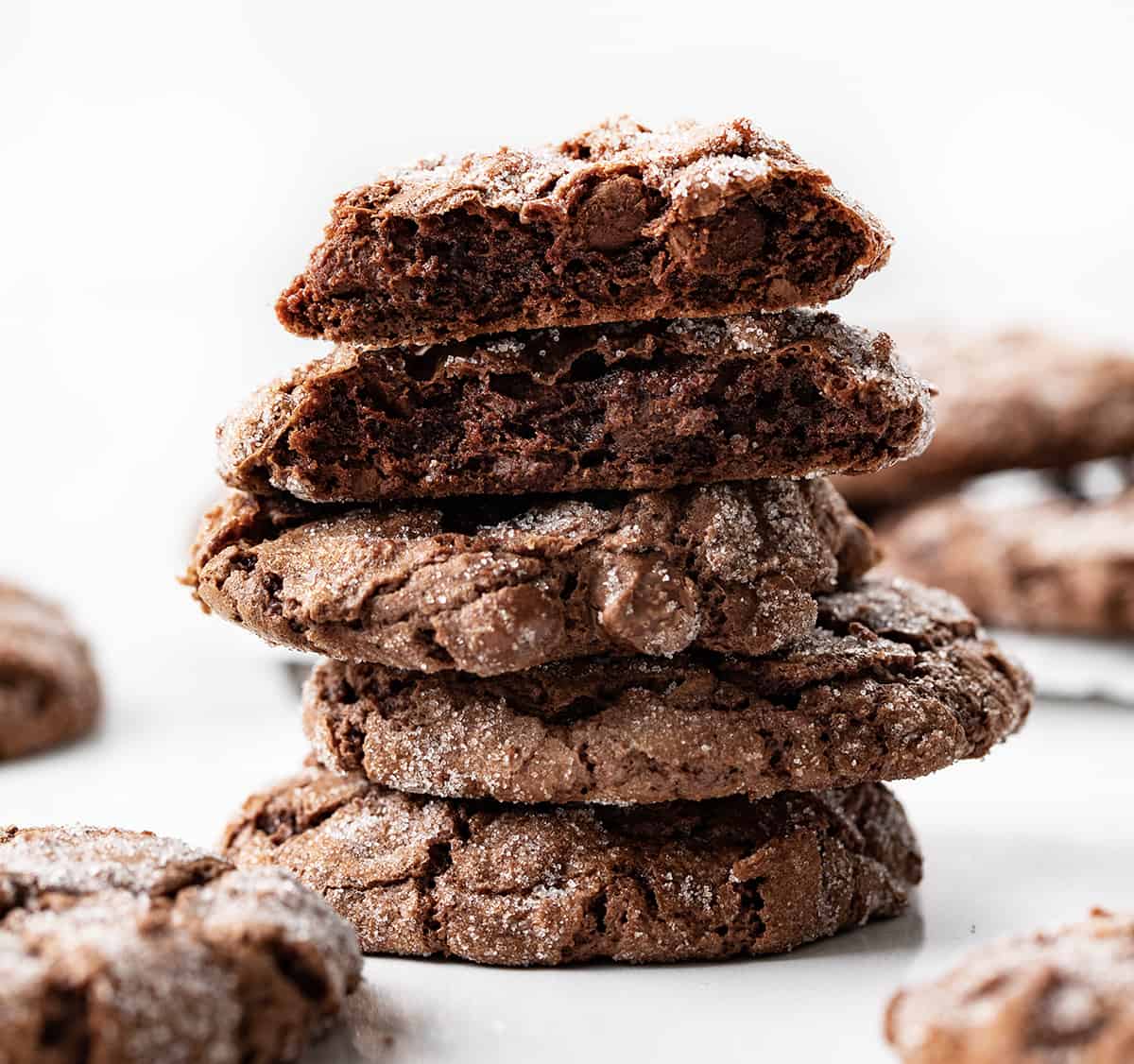 Dirty Chocolate Cookies are rich chocolate cookies rolled in sugar with a crispy outside and a soft, chewy center. Using two kinds of chocolate in the cookies makes them a decadent and indulgent treat with a deep chocolate flavor in every bite. They are worth the crumbs and sugar that may get everywhere! Dirty Chocolate Cookies on a White Counter with One Cookies Broken Showing the Inside Texture.