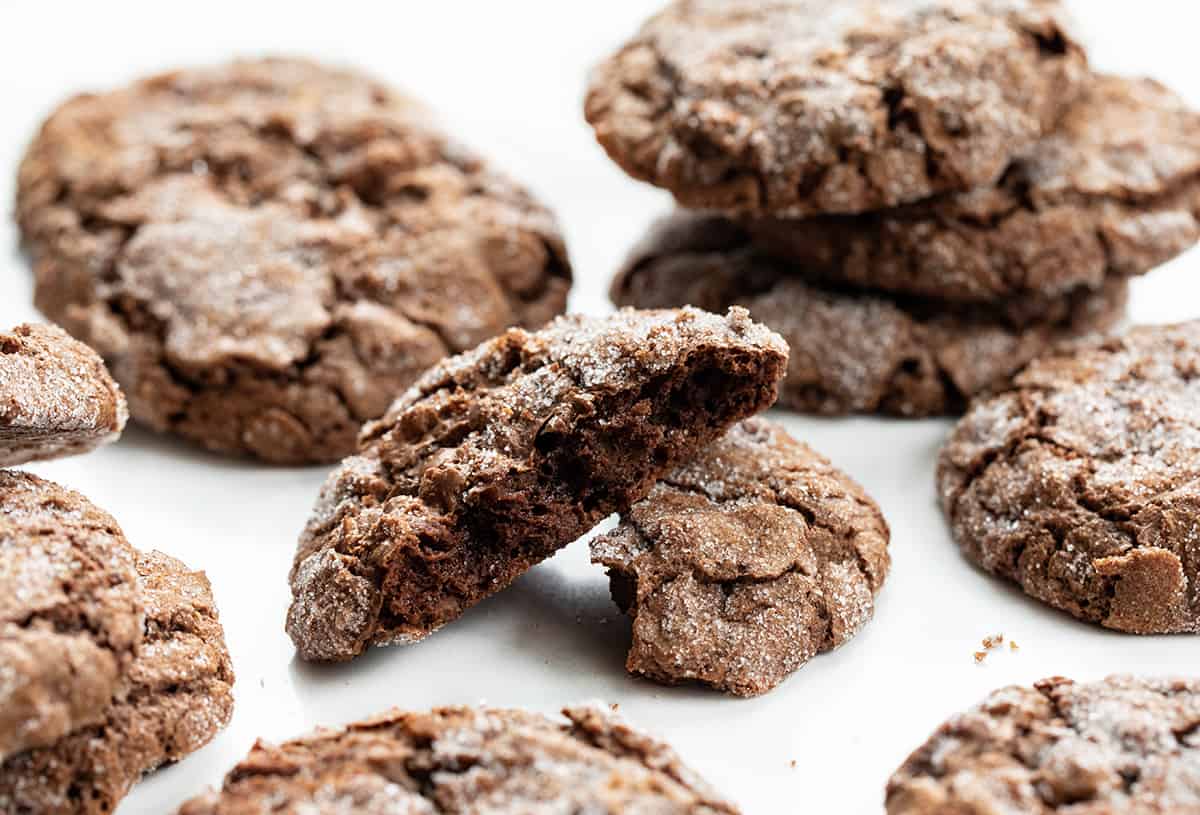 Dirty Chocolate Cookies are rich chocolate cookies rolled in sugar with a crispy outside and a soft, chewy center. Using two kinds of chocolate in the cookies makes them a decadent and indulgent treat with a deep chocolate flavor in every bite. They are worth the crumbs and sugar that may get everywhere! Dirty Chocolate Cookies on a White Counter with One Cookies Broken Showing the Inside Texture.