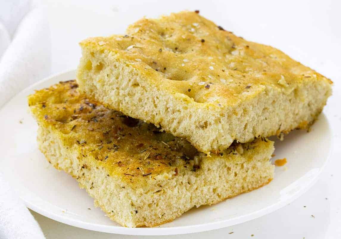 Focaccia Bread is an Italian bread that is made with lots of olive oil and full of Italian seasoning for a delicious and savory snack or appetizer. focaccia bread, bread recipes, Italian bread, i am baker, iambaker, homemade focaccia bread recipe, baking bread Easy Focaccia Bread Cut into Slices and on a White Plate
