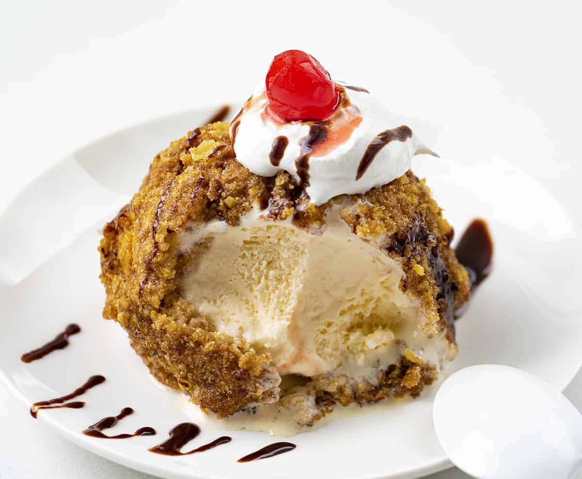 Fried Ice Cream is a frozen ball of ice cream, coated in egg whites, vanilla wafers, cornflakes, and cinnamon, deep-fried to a golden brown. Dessert, No Bake Dessert, Hot Dessert, Ice Cream, Ice Cream Dessert, Summer Desserts, recipes, air fryer fried ice cream, i am baker, iambaker Scoop of Fried Ice Cream on a Plate with Bites Removed and its Drizzled in Hot Fudge.