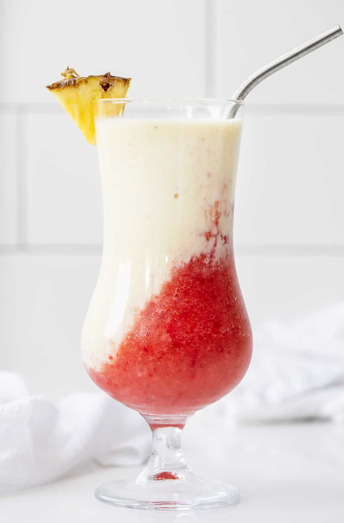 Lava Flow Drink is a tropical blended cocktail that combines the flavors of strawberries, pineapple, and coconut. It has a stunning visual appearance when the red layer flows into the creamy coconut layer, like an erupting volcano! One Lava Flow Drink on a White Counter.