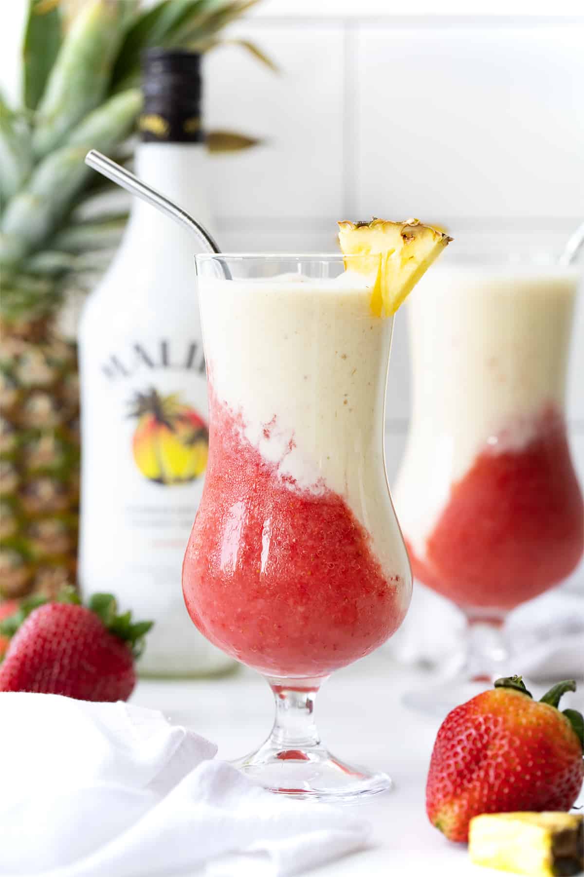 Lava Flow Drink is a tropical blended cocktail that combines the flavors of strawberries, pineapple, and coconut. It has a stunning visual appearance when the red layer flows into the creamy coconut layer, like an erupting volcano! The Lava Flow Drink on a Counter with Pineapple, Malibu, and another Lava Flow Behind It.