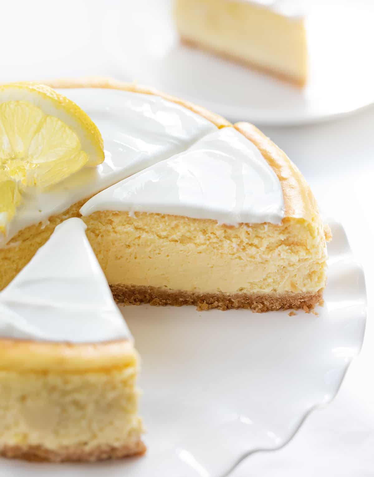 Lemon Cheesecake is a graham cracker crust filled with a light and smooth lemon-flavored filling, all topped with a creamy sour cream layer. If you appreciate the vibrant and refreshing taste of lemons and love cheesecake, this will be your new favorite dessert! One Lemon Cheesecake on a White Cake Stand with Ruffles and Pieces Cut with a Piece on a Plate in the Background.