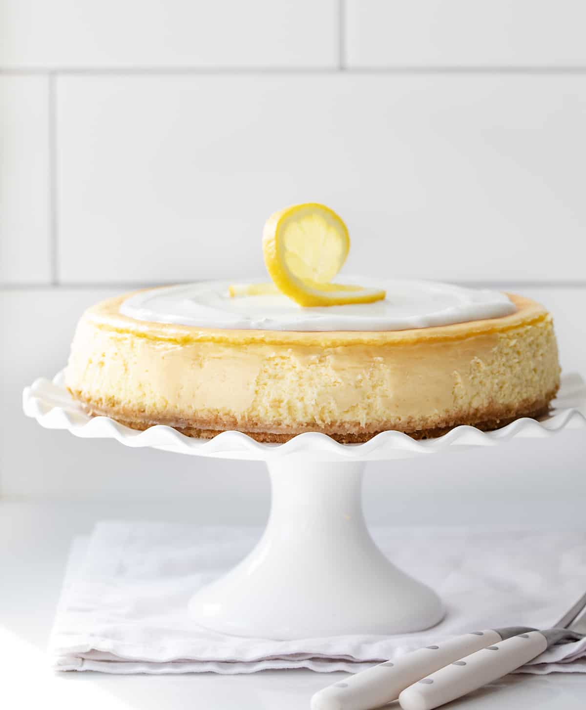 Lemon Cheesecake is a graham cracker crust filled with a light and smooth lemon-flavored filling, all topped with a creamy sour cream layer. If you appreciate the vibrant and refreshing taste of lemons and love cheesecake, this will be your new favorite dessert! Lemon Cheesecake on a White Cake Stand with a Ruffled Edge on a White Counter in Front of a Subway Tiled Wall.