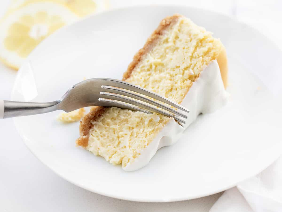 Lemon Cheesecake is a graham cracker crust filled with a light and smooth lemon-flavored filling, all topped with a creamy sour cream layer. If you appreciate the vibrant and refreshing taste of lemons and love cheesecake, this will be your new favorite dessert! Piece of Lemon Cheesecake on a White Plate with a Fork Taking a Bite.