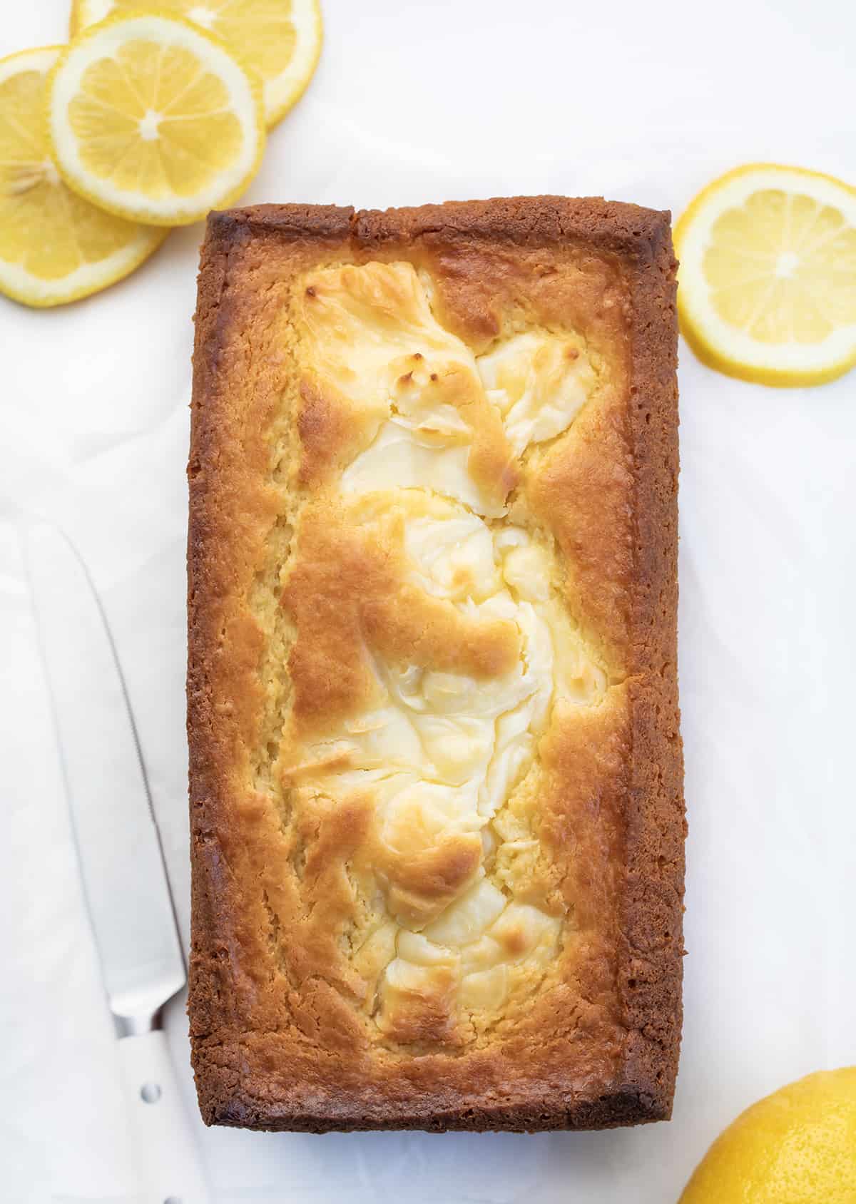 Lemon Cream Cheese Loaf is a moist and tender cake bursting with lemon flavor with a rich cream cheese swirl. It is delicious and visually stunning, with a beautiful marbled pattern from the swirl. It's a delightful option for breakfast, brunch, a sweet snack, or dessert! Overhead of a Lemon Cream Cheese Loaf on Counter with Lemons.