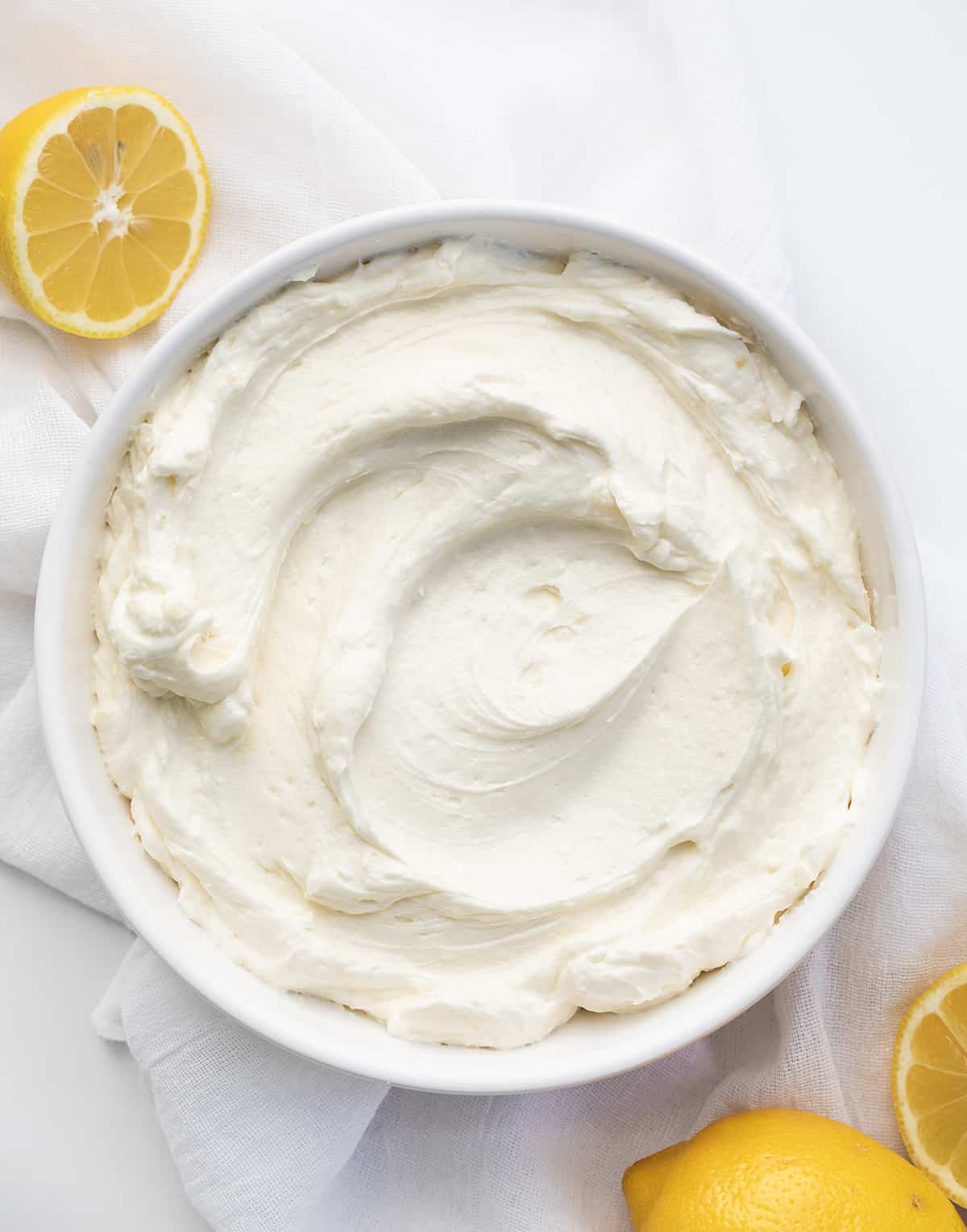Lemon Ermine Frosting is a smooth, creamy, and buttery frosting made with a cooked lemon juice and flour mixture for just the right lemony zing in every taste. It has a smooth-as-silk texture and a POP of bright lemon tartness that pairs so well with the subtle sweetness of classic ermine frosting. It will give many baked goods a bright lemon flavor! It is perfect for piping and frosting cakes! Bowl of Lemon Ermine Frosting Smoothed Out with Lemons around.
