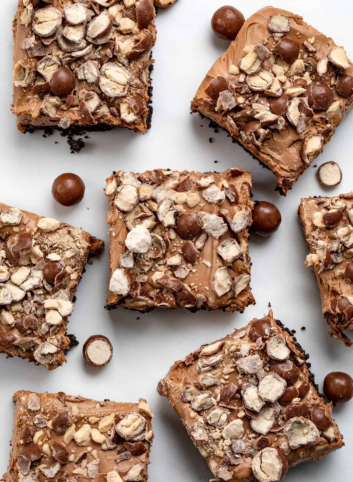 Chocolate Malt Shoppe Bars are creamy chocolatey bars with added chocolate malt powder over an Oreo cookie crust and topped with crushed malted milk ball (whopper) candies. This no-bake treat is like enjoying your favorite chocolate malted milkshake but in a dessert bar without any ice cream. Let each bite take you back to the nostalgic charm of your favorite malt shop! Chocolate Malt Shoppe Bars on a White Counter from Overhead.