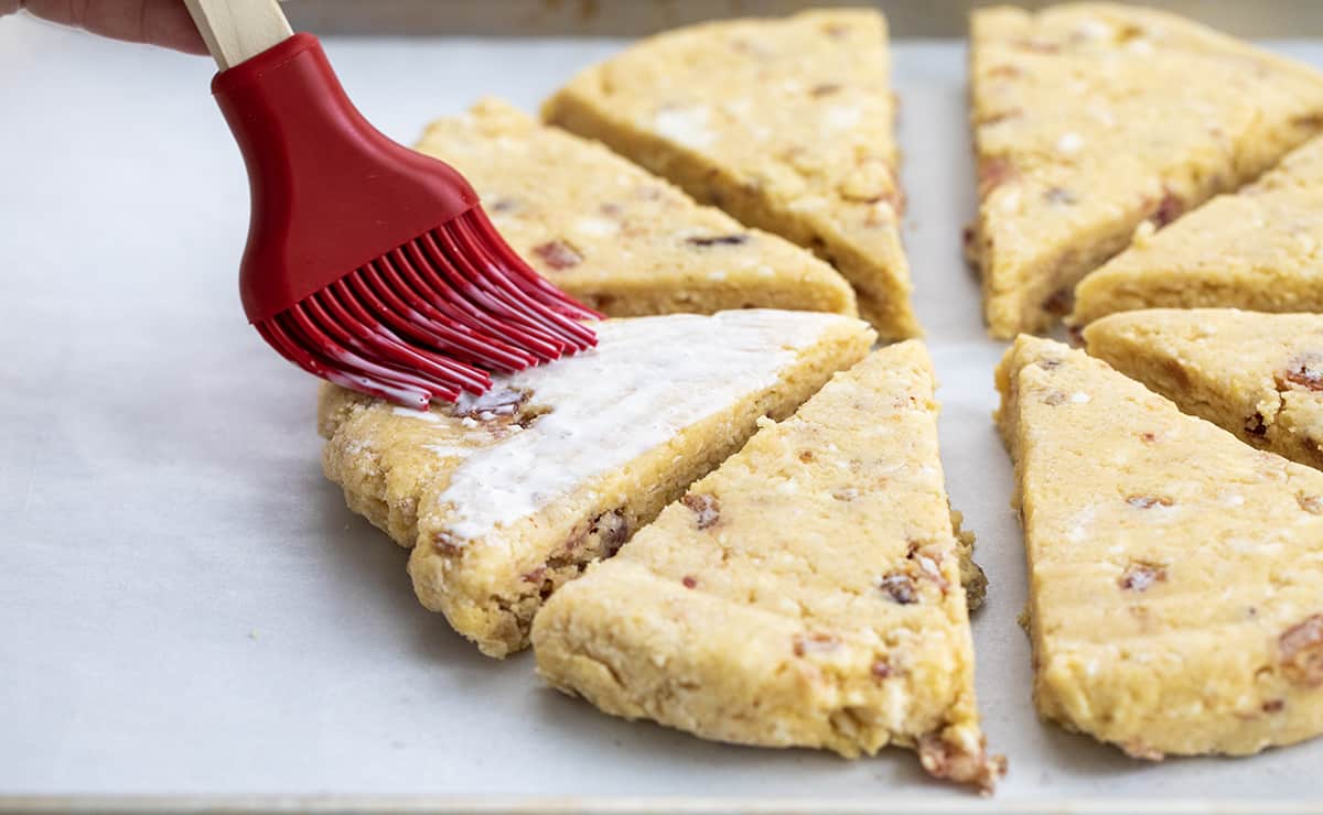This Maple Bacon Scones recipe is a unique twist on a classic breakfast treat, featuring the perfect blend of sweet maple syrup and salty bacon. The scone has a tender inside and crispy top and is finished with a delicious maple glaze and crumbled bacon. This recipe is easy to follow and uses common ingredients, so don't be intimidated by the thought of making scones! Brushing Milk Over Chilled Maple Bacon Scones Before Baking.