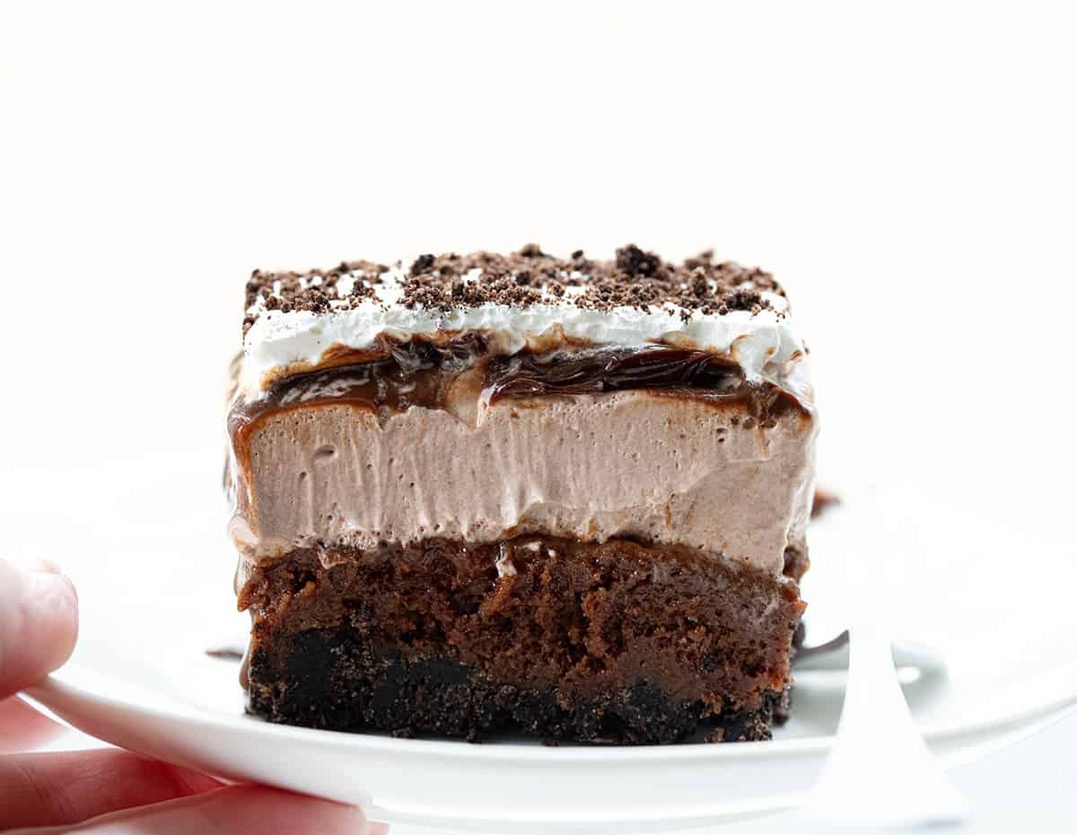 Chocolate Mud Bars are a rich and indulgent layered dessert with an Oreo cookie crust topped with fudgy brownies, chocolate mousse, hot fudge, and whipped cream. It's a chocolate lover's dream dessert that combines different textures and flavors to create a truly lavish treat! Hand Holding a Plate with Chocolate Mud Bar.