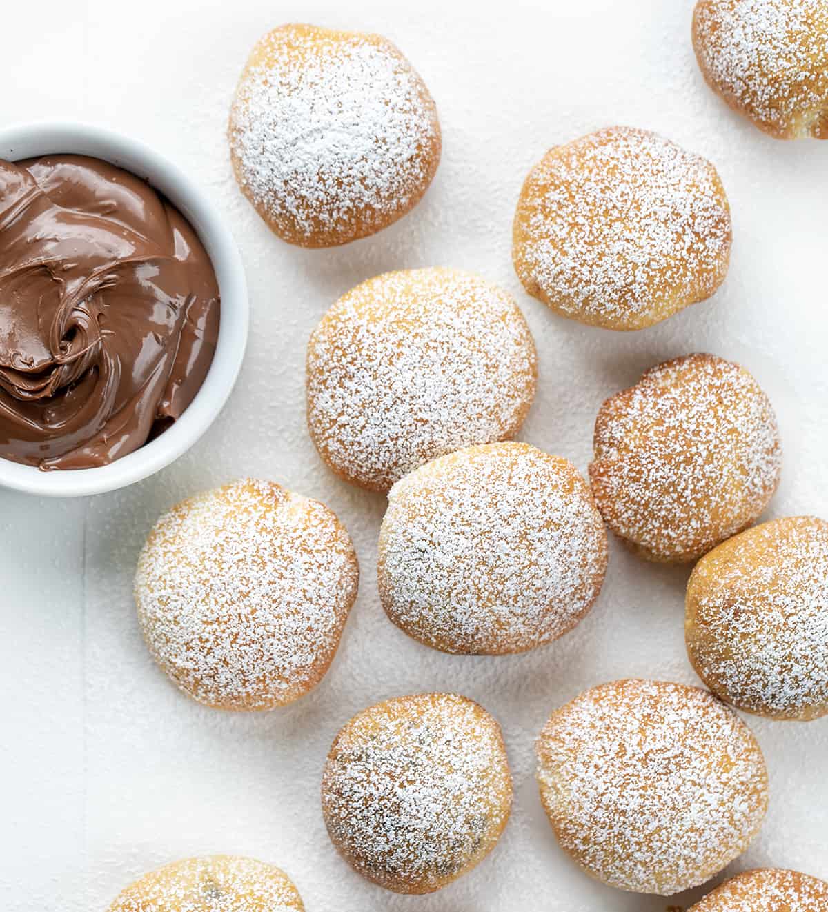Nutella Bombs are sweet treats made with just four simple ingredients and cooked in an air fryer. They start with a can of refrigerated biscuits that are separated and filled with Nutella. After rolled up into balls, they are cooked to a golden brown. Then, to top it all off, literally, confectioners' sugar is dusted over the tops. Each bite is bursting with Nutella! (I will also let you know how to bake these in the oven. Nutella Bombs on a White Counter and Dusted with Confectioners Sugar Next to a Bowl of Nutella.