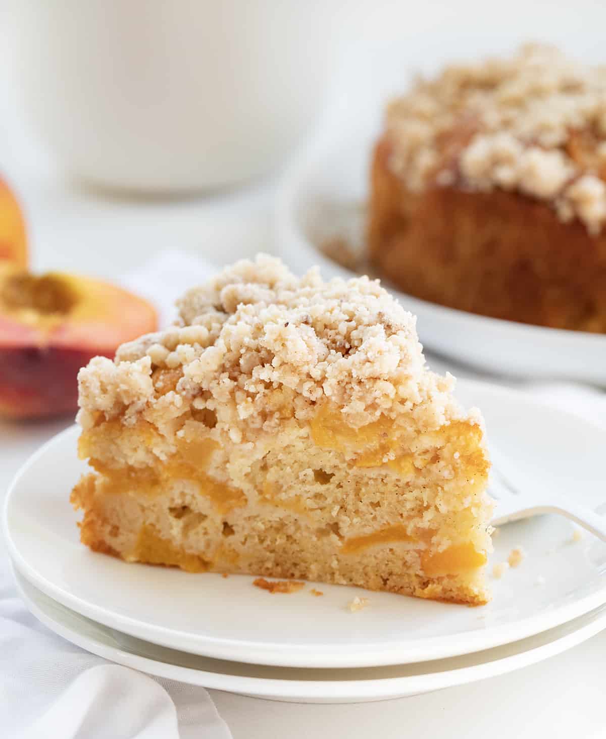 Peach Cake is loaded with peaches, baked to a tender and moist crumb, then drenched with a sweet warm milk mixture and sprinkled with a crumble topping. This is an absolute must-try for anyone who loves peaches or is simply craving a sweet dessert! Peach Cake on a Plate with the Whole Cake Behind it and Peaches.
