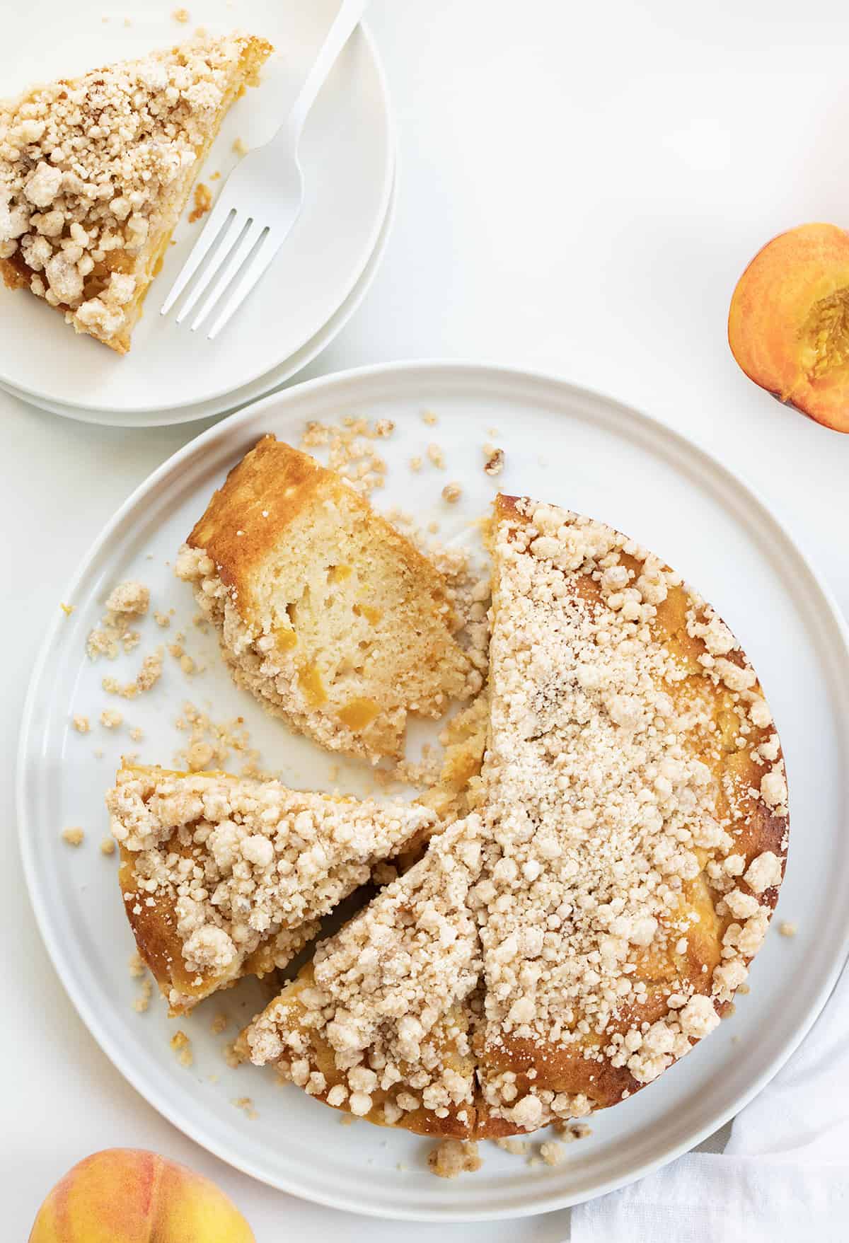 Peach Cake is loaded with peaches, baked to a tender and moist crumb, then drenched with a sweet warm milk mixture and sprinkled with a crumble topping. This is an absolute must-try for anyone who loves peaches or is simply craving a sweet dessert! Whole Peach Cake on White Platter With Half of the Pieces Cut and One on its Side.
