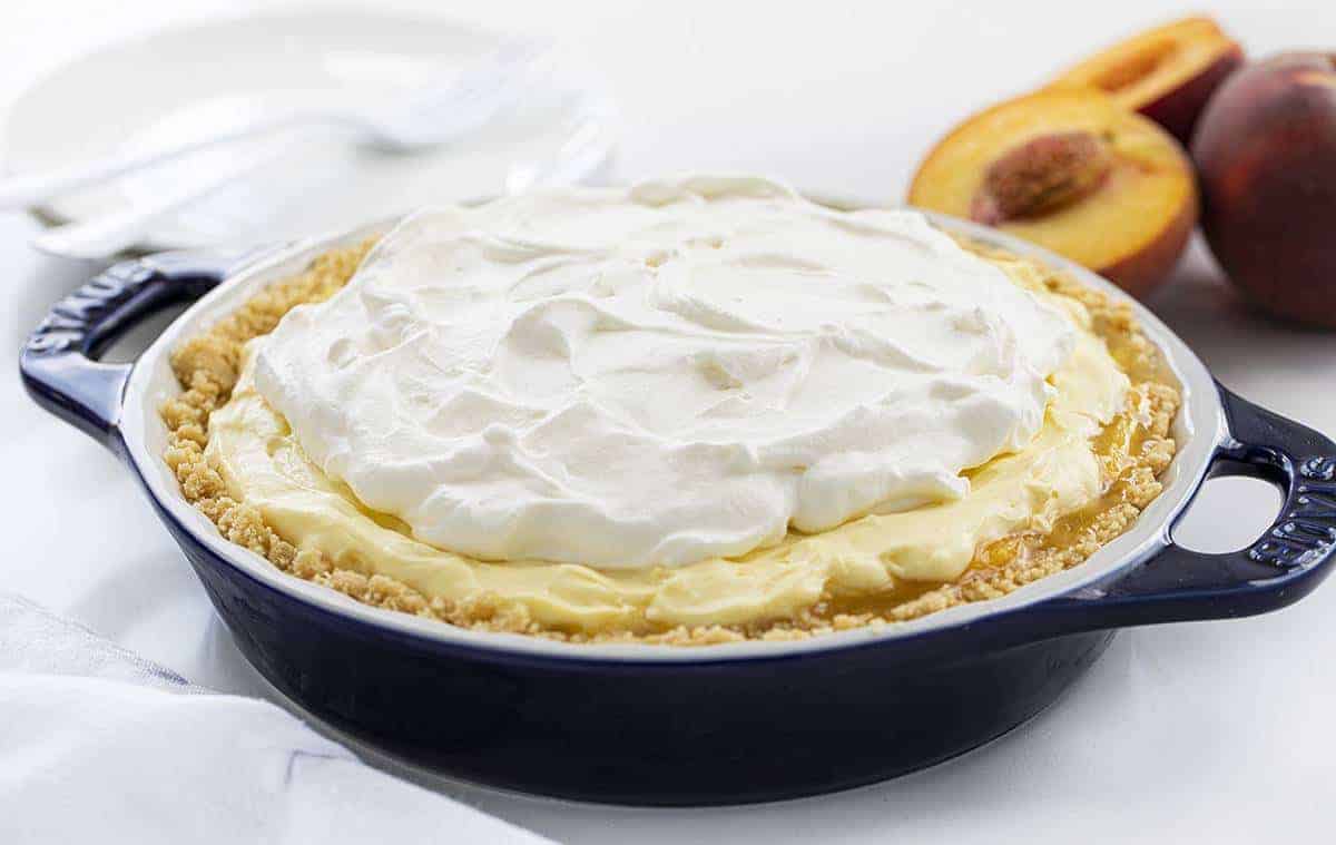 The best no-bake desserts to get your sweet tooth through the summer! no bake, no bake desserts, desserts, recipes, cheesecake, cookies, bars, cake, pudding, mousse, puppy chow, muddie buddies, rice krispy bars, dessert, memorial day dessert, labor day desserts, i am baker, iambaker Peaches and Cream Pie in a Blue Pie Dish with Peaches in Back.