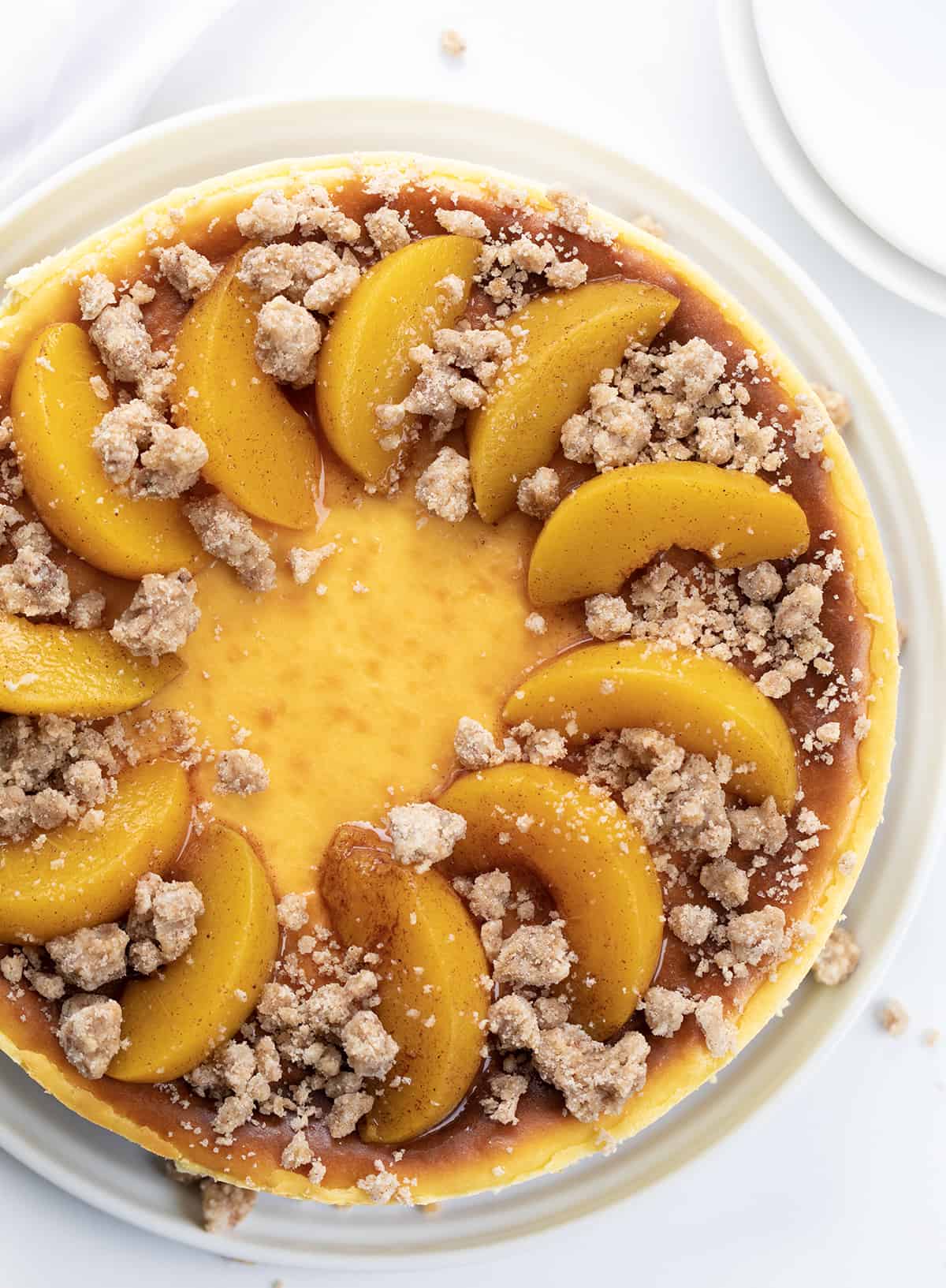 Peach Crumble Cheesecake is a buttery graham cracker crust topped with layers of cheesecake, crumbly streusel, and peaches! You will enjoy the creaminess of the cheesecake with the juicy peaches and the streusel texture in each delectable bite. Looking Down on a Peach Crumble Cheesecake on a White Counter with Towel and Plates.