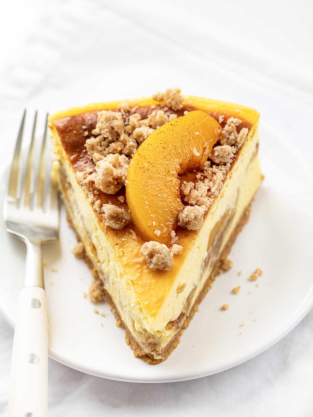 Peach Crumble Cheesecake is a buttery graham cracker crust topped with layers of cheesecake, crumbly streusel, and peaches! You will enjoy the creaminess of the cheesecake with the juicy peaches and the streusel texture in each delectable bite. Slice of Peach Crumble Cheesecake on a White Plate with a White Fork.