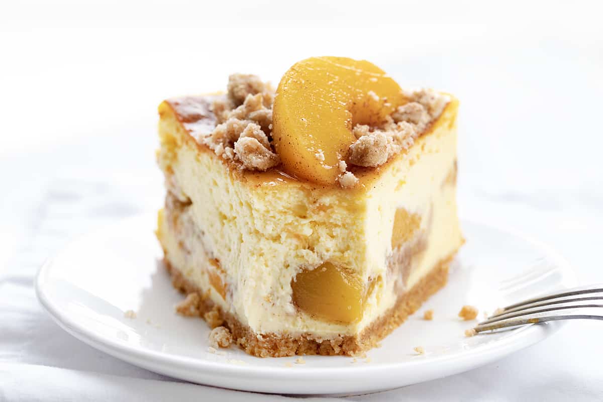 Peach Crumble Cheesecake is a buttery graham cracker crust topped with layers of cheesecake, crumbly streusel, and peaches! You will enjoy the creaminess of the cheesecake with the juicy peaches and the streusel texture in each delectable bite. Piece of Peach Crumble Cheesecake on a White Plate with a Bite Removed.