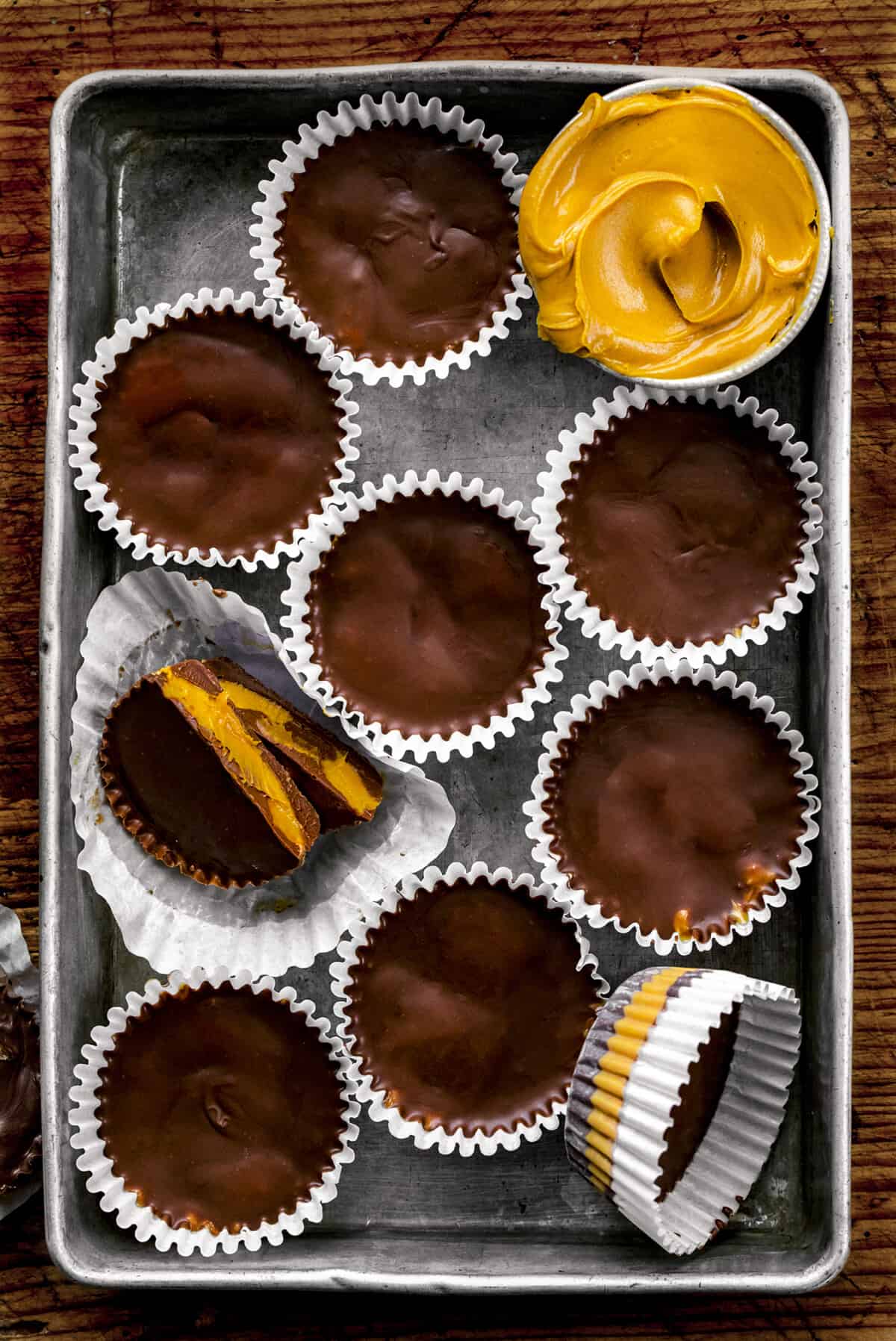 Homemade Peanut Butter Cups... SO YUM! I am a sucker for everything chocolate and peanut butter. And, when I can make my own treats with this flavor combination, it is even better! With this treat, there are no added preservatives like when buying peanut butter cups from the store. If you need some holiday treats, add them to your easy-finger food Christmas party options or as a part of your Christmas dessert charcuterie board! Homemade Peanut Butter Cups in a Pan with Some Extra Peanut Butter and One Cut in Half.