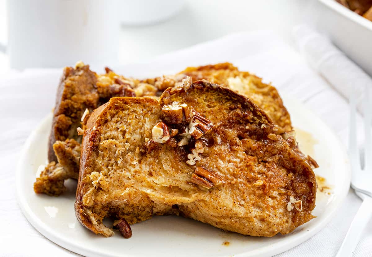 Pumpkin French Toast Casserole is slices of Challah bread dipped into a pumpkin-flavored custard mixture, sprinkled with pecans, and baked for a warm breakfast or brunch. It's a perfect recipe to prepare the night before and get in the oven the next morning! Pieces of Pumpkin French Toast on a White Plate with Coffee in Background.