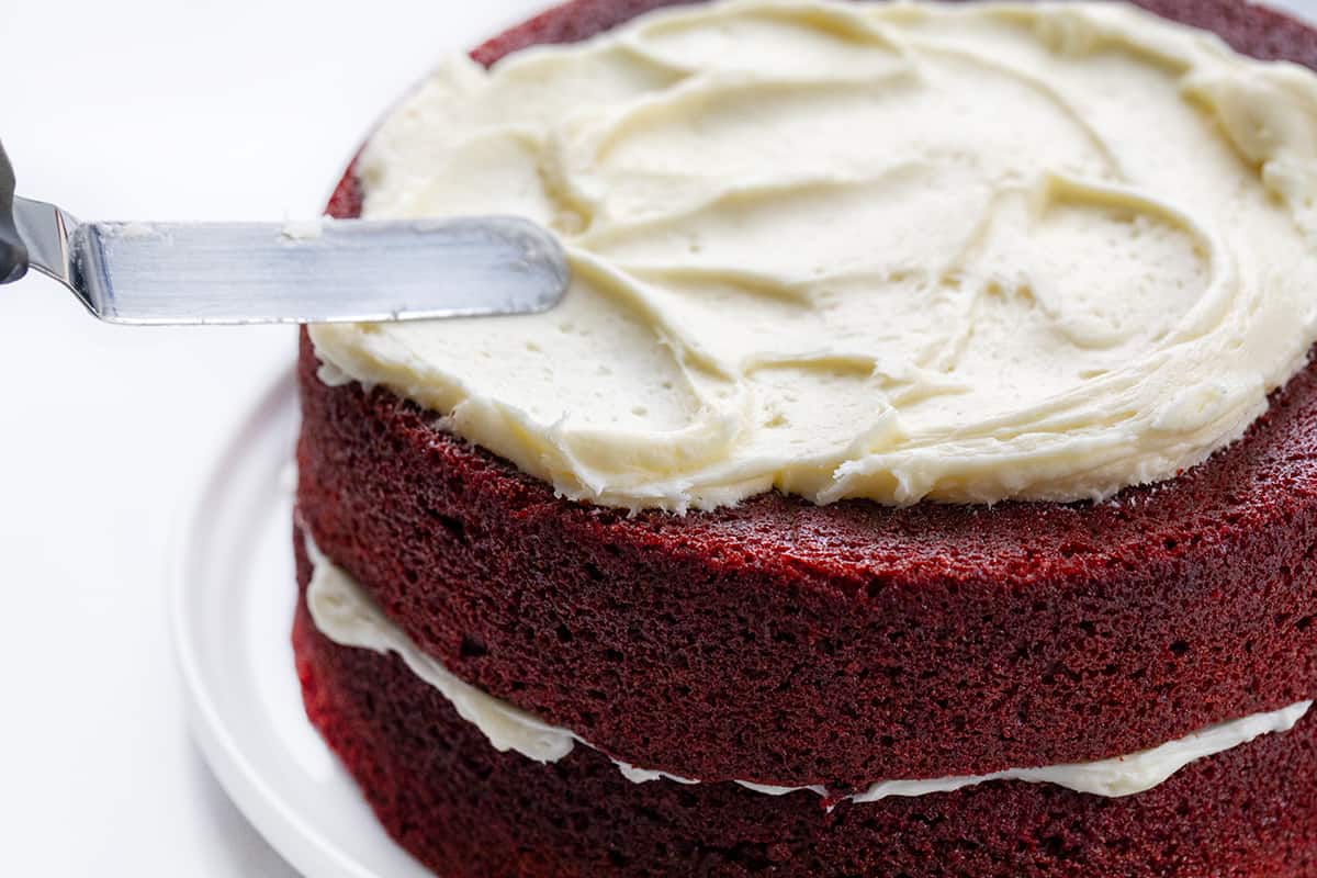 The perfect Red Velvet cake has a rich, deep flavor and a color to match! Then paired with the perfect cream cheese frosting, this just might be the perfect cake! Small offset spatula smoothing cream cheese frosting over layered red velvet cake.