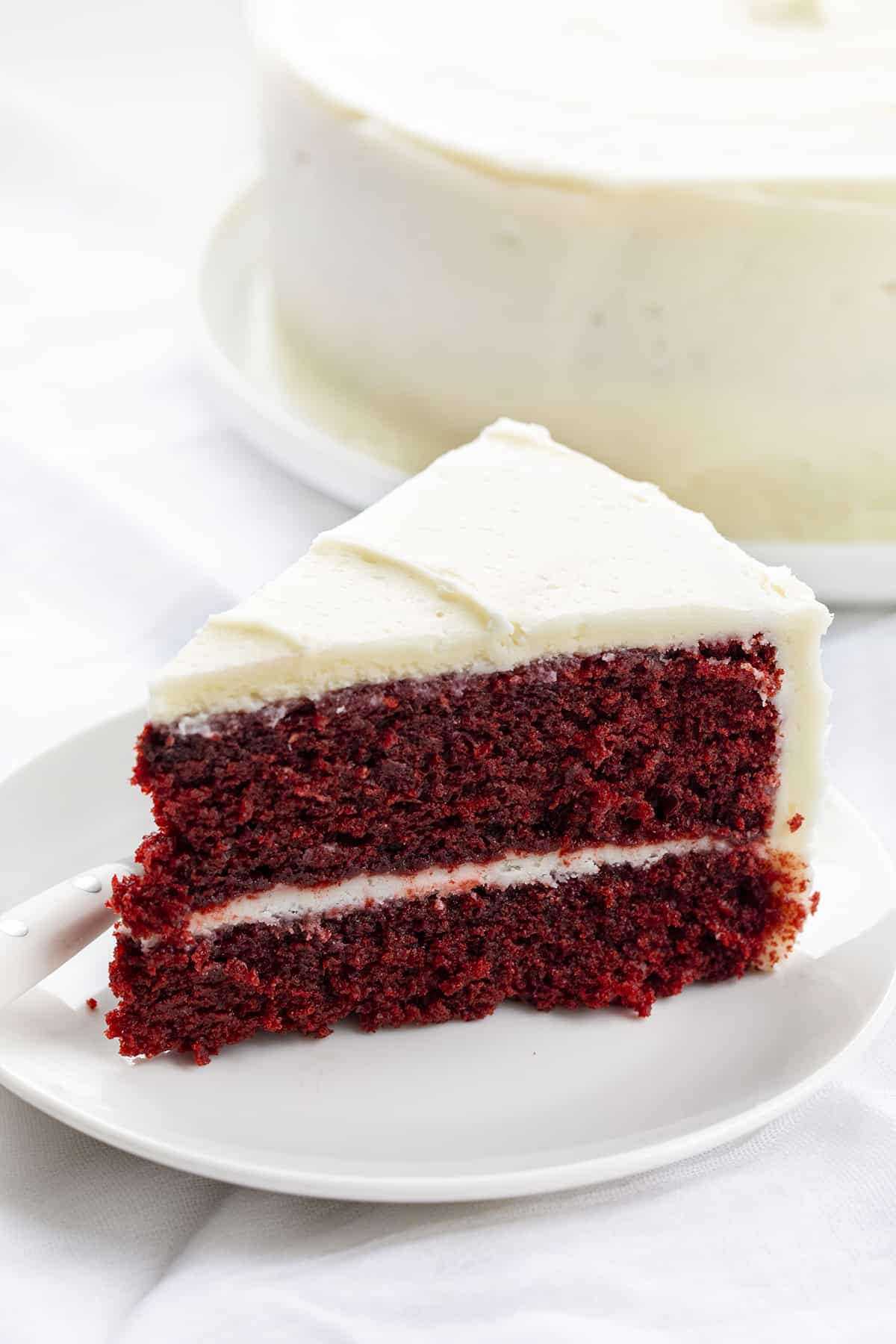 The perfect Red Velvet cake has a rich, deep flavor and a color to match! Then paired with the perfect cream cheese frosting, this just might be the perfect cake! Piece of Red velvet cake on a plate with the whole cake behind it in the background.