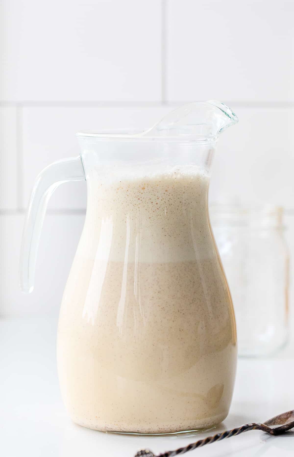If you love a smooth, creamy cocktail... this is the recipe for you! RumChata is described as a Caribbean rum that blends with Mexican spices and cream. It is the perfect addition to many cocktails and smart to have on hand during the holidays! Perfect for Christmas Cocktails, New Years Eve Cocktails, or even your company Christmas Party! Pitcher of Homemade Rum Chata with froth neat the top of the glass pitcher on a white counter.