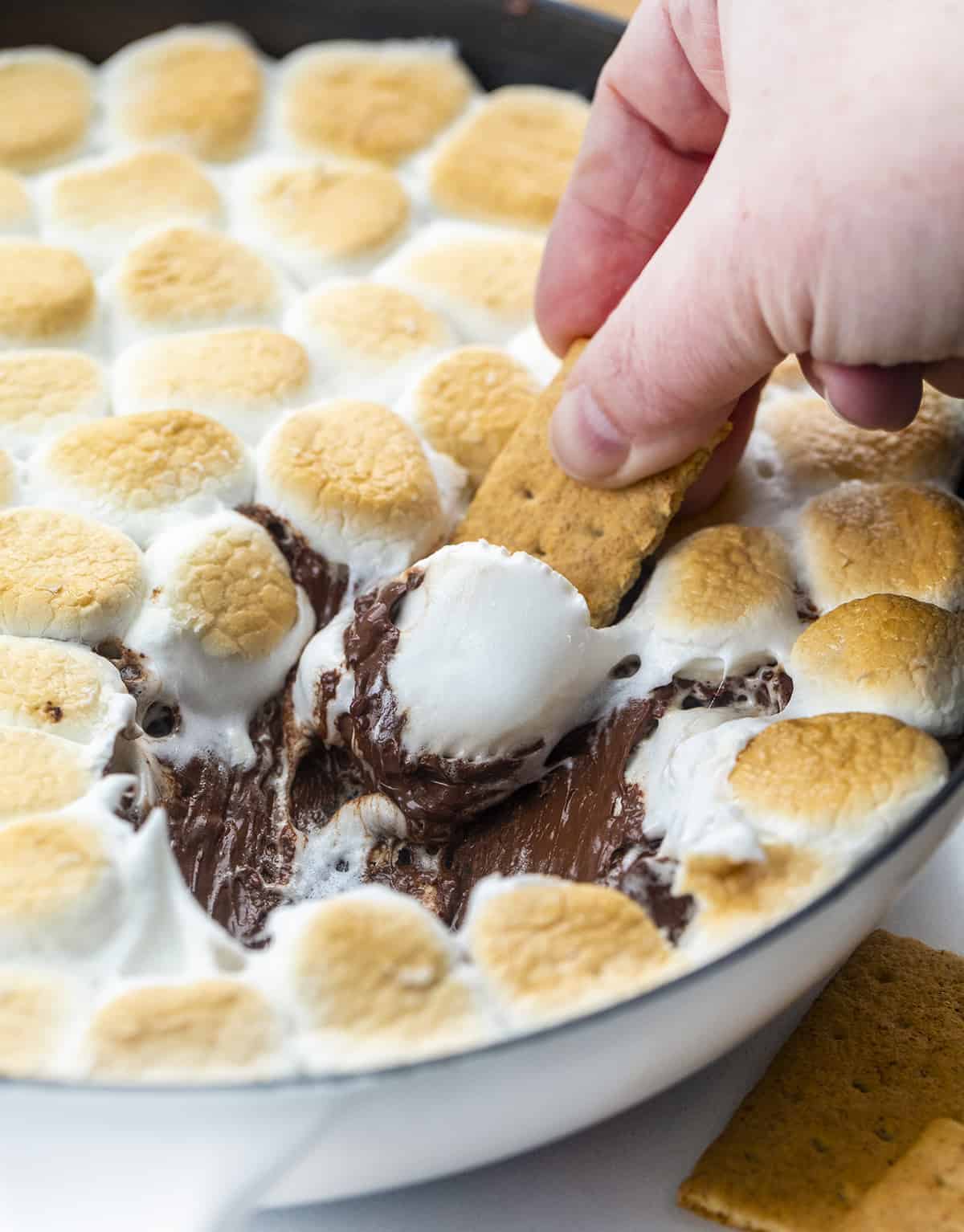 S'mores Dip is a warm, ooey-gooey, chocolatey dip made with melted chocolate chips that are topped with marshmallows and toasted in the oven. Appetizer, Dessert Appetizer, Smores Dip, Smores Dip in the Oven, Dessert, Chocolate, Marshmallow Dip, Skillet Desserts, Skillet Recipes, i am baker, iambaker Hand Dipping Graham Cracker into Smores Dip