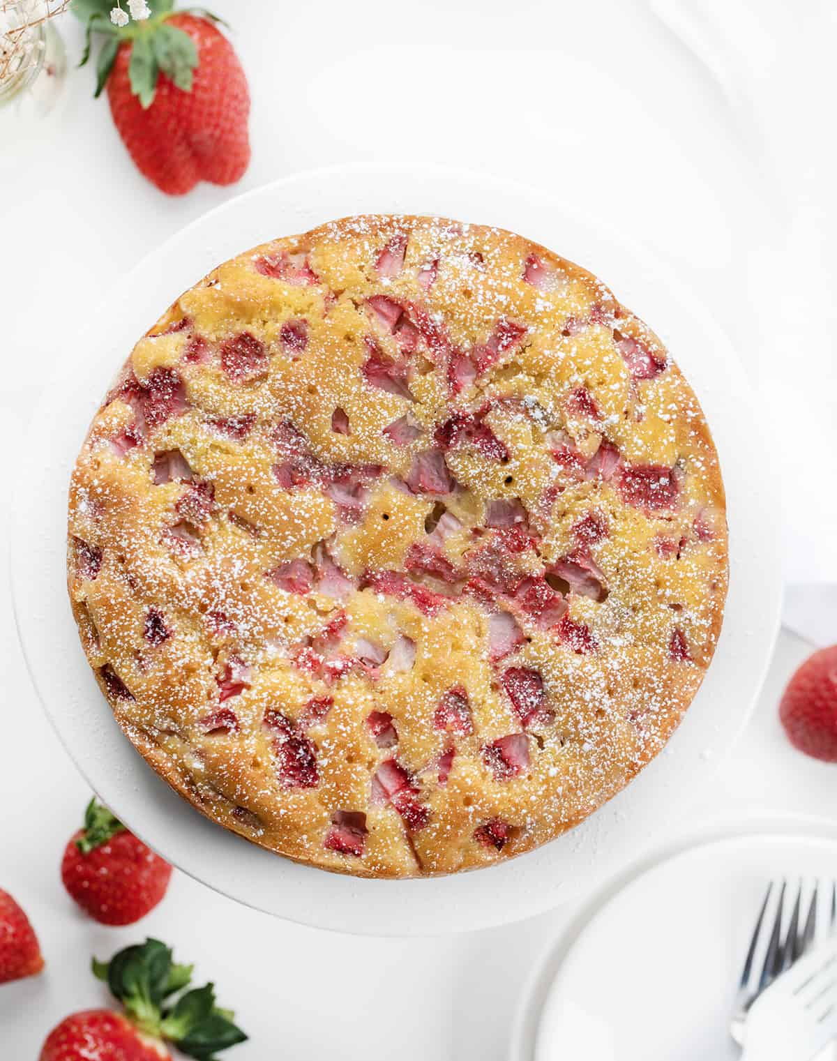 THE BEST STRAWBERRY CAKE YOU'LL EVER MAKE! This Fresh Strawberry Cake is a moist and fluffy cake filled and topped with fresh strawberries, baked to perfection, drenched in a sweet warm milk mixture, and dusted with confectioners' sugar. It's a refreshing and delightful treat! Whole Fresh Strawberry Cake on a White Plate with Strawberries in the Background.
