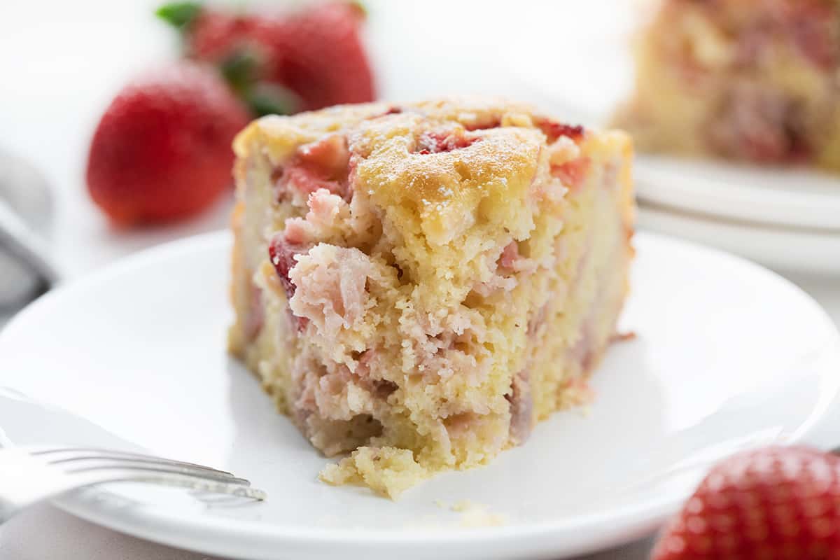 THE BEST STRAWBERRY CAKE YOU'LL EVER MAKE! This Fresh Strawberry Cake is a moist and fluffy cake filled and topped with fresh strawberries, baked to perfection, drenched in a sweet warm milk mixture, and dusted with confectioners' sugar. It's a refreshing and delightful treat! Piece of Fresh Strawberry Cake on a White Plate with Strawberries in the Background.