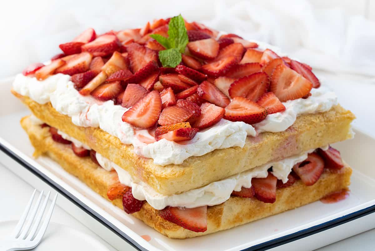 Sheet Pan Strawberry Shortcake is an elegant (but easy!) dessert that starts with the perfect white sheet cake cut in half and layered with fresh strawberries and a whipped cream cheese topping. It is a delicious and refreshing variation of the classic dessert perfect for summertime or any occasion! Sheet Pan Strawberry Shortcake in a Pan on a White Counter Turned at an Angle.