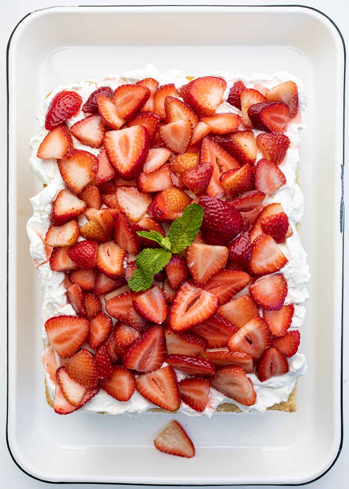 Sheet Pan Strawberry Shortcake is an elegant (but easy!) dessert that starts with the perfect white sheet cake cut in half and layered with fresh strawberries and a whipped cream cheese topping. It is a delicious and refreshing variation of the classic dessert perfect for summertime or any occasion! Looking Down onto a Sheet Pan Strawberry Shortcake with a Sprig of Mint in the Middle.