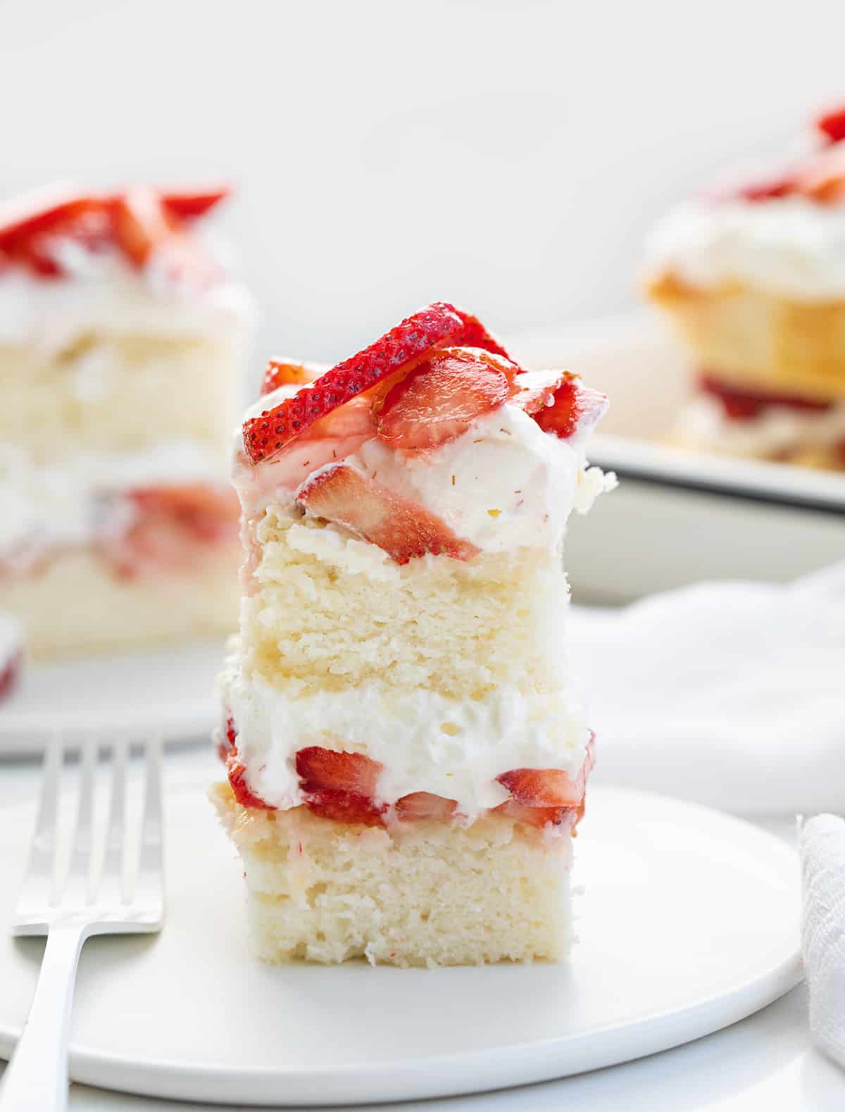 Sheet Pan Strawberry Shortcake is an elegant (but easy!) dessert that starts with the perfect white sheet cake cut in half and layered with fresh strawberries and a whipped cream cheese topping. It is a delicious and refreshing variation of the classic dessert perfect for summertime or any occasion! Slices of Sheet Pan Strawberry Shortcake on a White Counter with White Plates and Silverware.