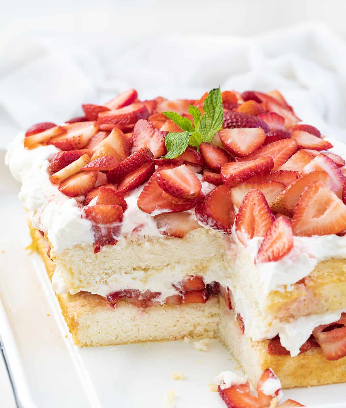 Sheet Pan Strawberry Shortcake is an elegant (but easy!) dessert that starts with the perfect white sheet cake cut in half and layered with fresh strawberries and a whipped cream cheese topping. It is a delicious and refreshing variation of the classic dessert perfect for summertime or any occasion! Cut Into Sheet Pan Strawberry Shortcake on a White Counter.