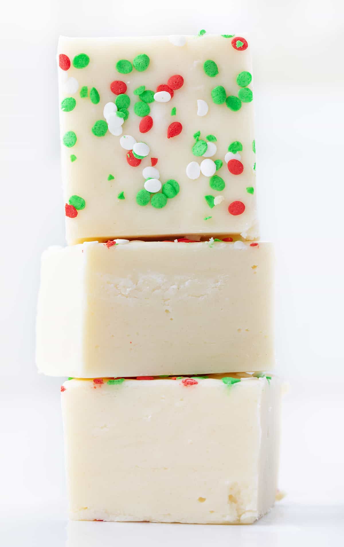 Sugar Cookie Fudge for the win! With sugar cookies being all the rage over the holidays, it's fun to add some variations to the popular cookie. This sugar cookie fudge is super simple to make, and with the added sprinkles, it can be transformed into a colorful holiday treat. Add a few squares to your Christmas dessert charcuterie board, serve it at a New Year's Eve party, or simply enjoy it as a sweet treat. Stack of Pieces of Sugar Cookie Fudge on a White Counter.