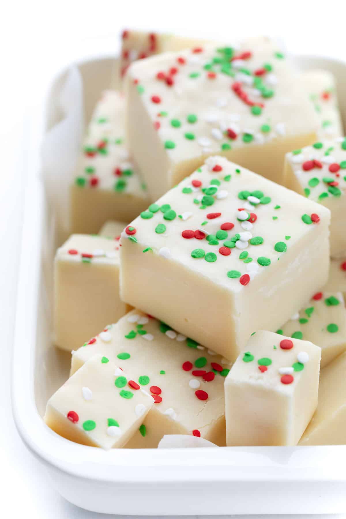 Sugar Cookie Fudge for the win! With sugar cookies being all the rage over the holidays, it's fun to add some variations to the popular cookie. This sugar cookie fudge is super simple to make, and with the added sprinkles, it can be transformed into a colorful holiday treat. Add a few squares to your Christmas dessert charcuterie board, serve it at a New Year's Eve party, or simply enjoy it as a sweet treat. Pieces of Sugar Cookie Fudge that has green and red sprinkles in a dish.