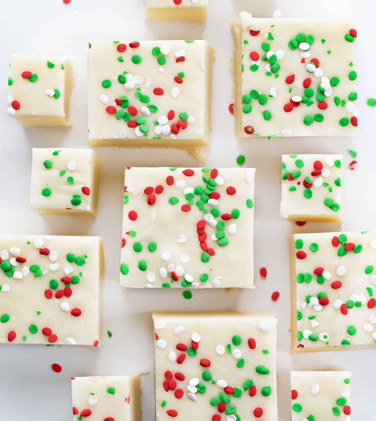 Sugar Cookie Fudge for the win! With sugar cookies being all the rage over the holidays, it's fun to add some variations to the popular cookie. This sugar cookie fudge is super simple to make, and with the added sprinkles, it can be transformed into a colorful holiday treat. Add a few squares to your Christmas dessert charcuterie board, serve it at a New Year's Eve party, or simply enjoy it as a sweet treat. Pieces of Sugar Cookie Fudge on a White COunter with Green and Red Spinkles.