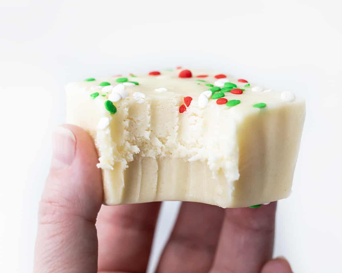 Sugar Cookie Fudge for the win! With sugar cookies being all the rage over the holidays, it's fun to add some variations to the popular cookie. This sugar cookie fudge is super simple to make, and with the added sprinkles, it can be transformed into a colorful holiday treat. Add a few squares to your Christmas dessert charcuterie board, serve it at a New Year's Eve party, or simply enjoy it as a sweet treat. Hand Holding a Piece of Sugar Cookie Fudge with a Bite Taken Out.