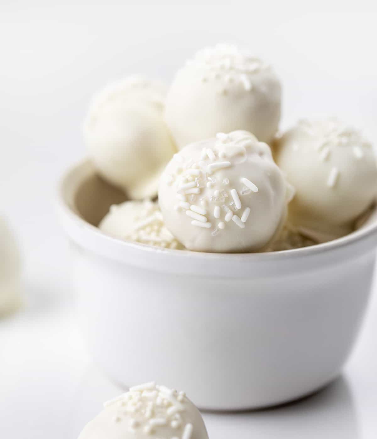 Sugar Cookie Truffles are a sweet treat made with crumbled sugar cookies and cream cheese, coated in white chocolate, and topped with sprinkles. They have all of the flavors you love from a sugar cookie packed into a decadent bite-sized treat. This three-ingredient recipe will be a perfect addition to your Christmas dessert charcuterie board! Sugar Cookie Truffles in a White Bowl on a White Counter.