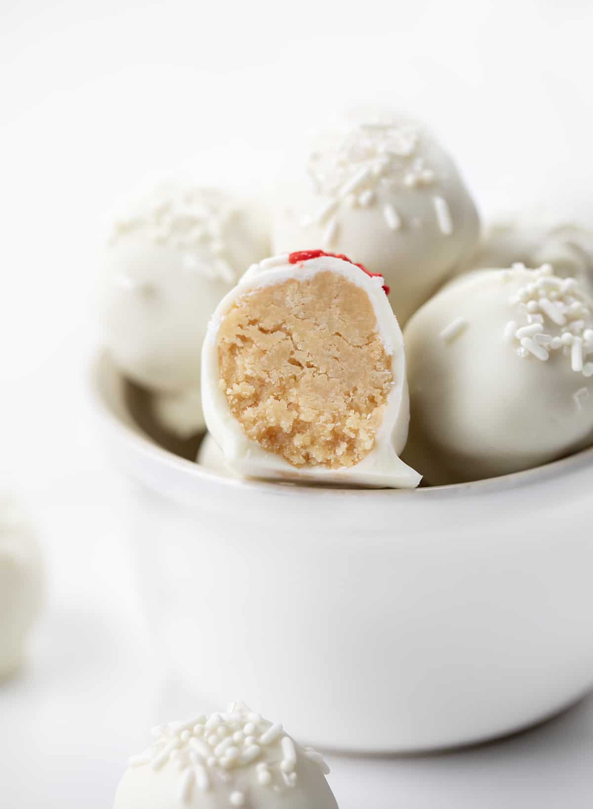 Sugar Cookie Truffles are a sweet treat made with crumbled sugar cookies and cream cheese, coated in white chocolate, and topped with sprinkles. They have all of the flavors you love from a sugar cookie packed into a decadent bite-sized treat. This three-ingredient recipe will be a perfect addition to your Christmas dessert charcuterie board! One cut in half Sugar Cookie Truffles on a Stack But Showing the Inside Texture.