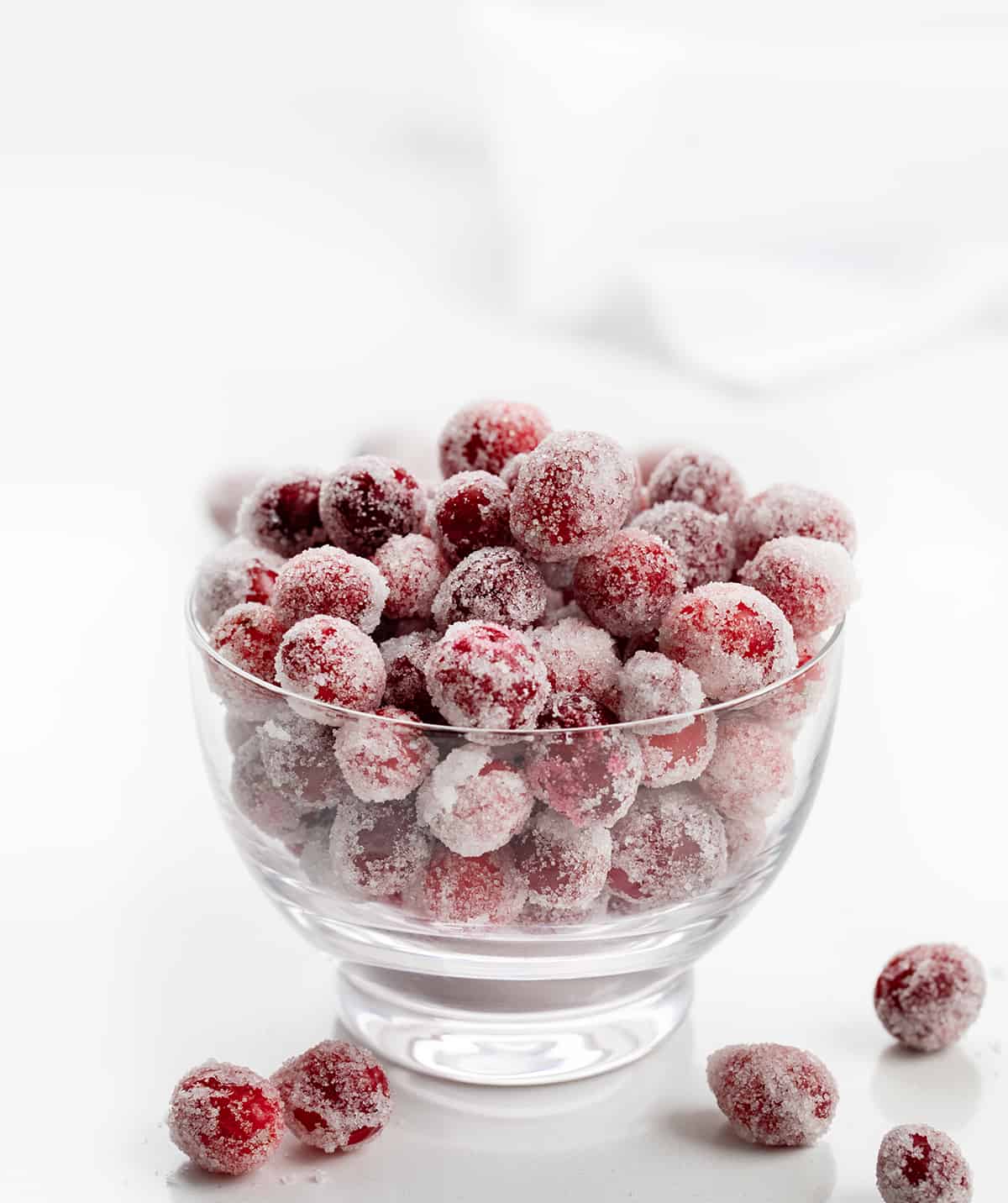 Sugared Cranberries are fresh cranberries coated in simple syrup and tossed in sugar for the perfect festive treat. The best part about this recipe is that it is only three simple ingredients--cranberries, sugar, and water! These are ideal for Cranberry Punch Recipes, Christmas Desserts, a Holiday Fruit Tray, or any Cocktail or Mocktail recipes! Bowl of Sugared Cranberries on a White Counter.