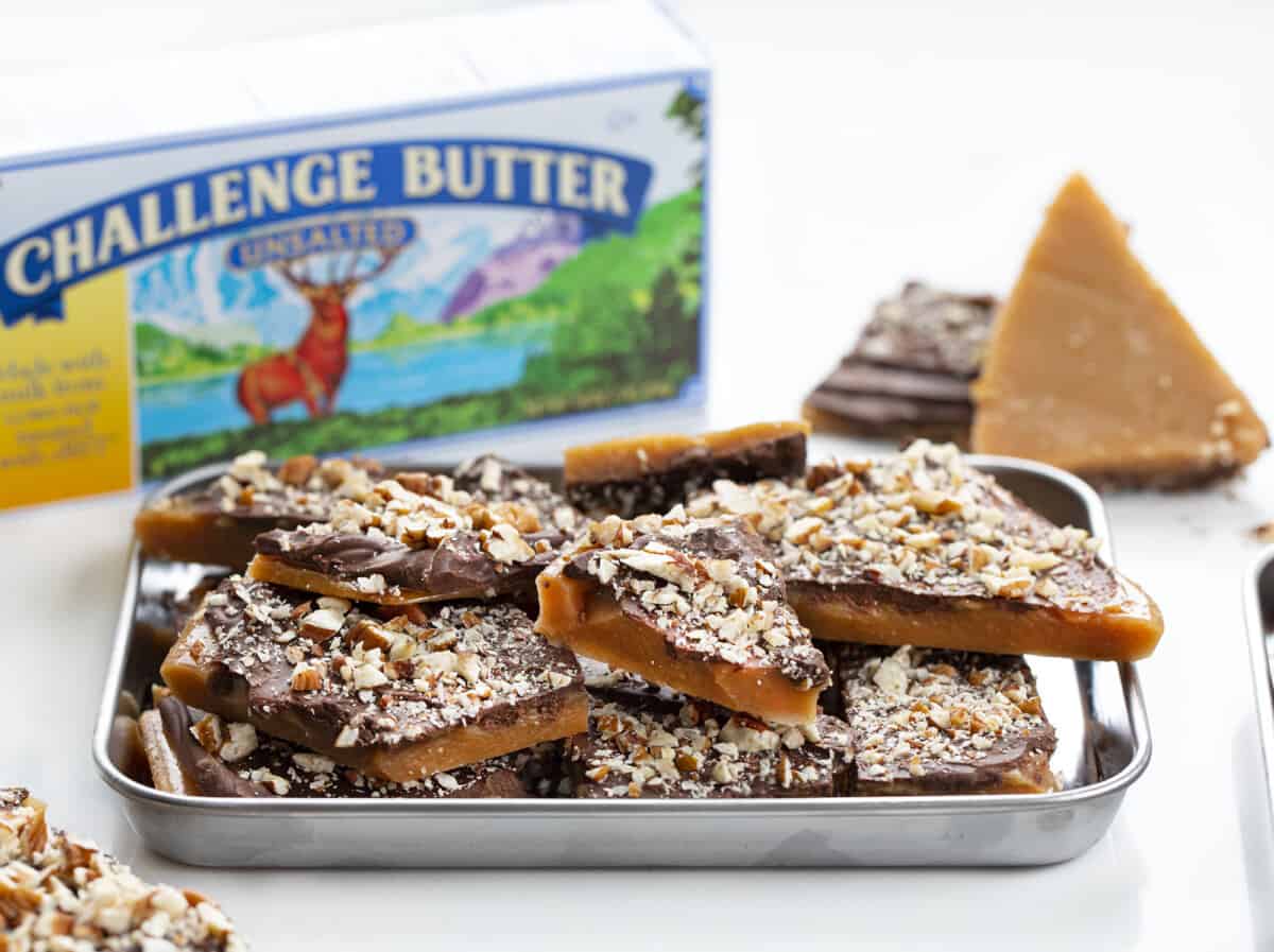 Learn how to make the BEST English Toffee that looks and tastes amazing! Rich buttery toffee covered with milk chocolate and chopped pecans. Just 4 ingredients! English toffee is the perfect holiday candy treat for parties and Christmas gifts. Tray of English Toffee Broken into Pieces and a Box of Challenge Butter Behind it.